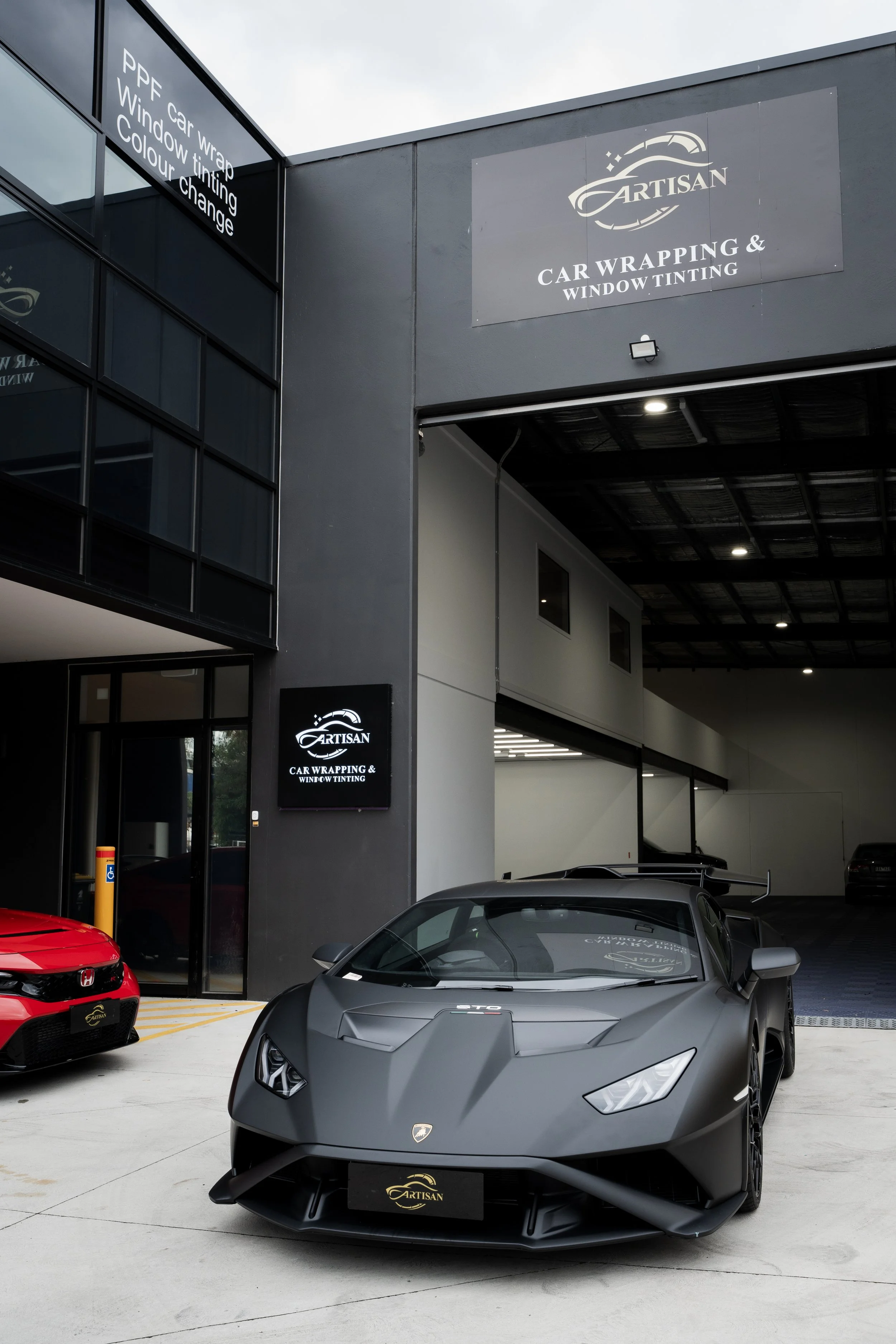 A sleek, matte black Lamborghini sports car parked outside an auto detailing shop called Artisan, which specializes in car wrapping and window tinting. The shop's signage is visible on the building and on a digital sign near the entrance.