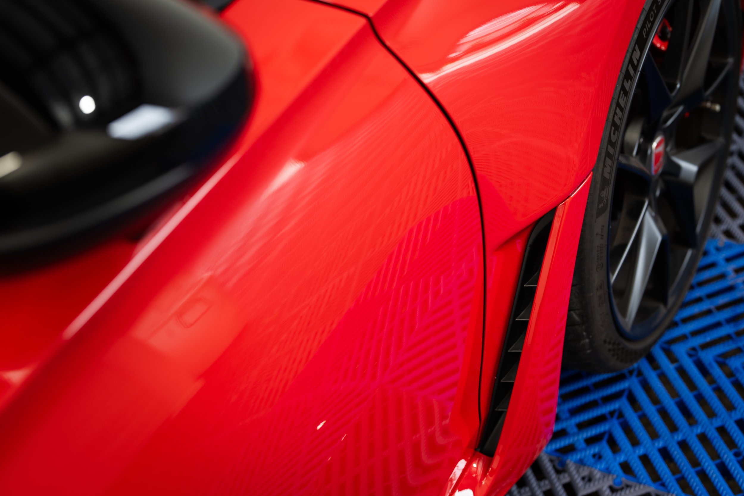 Close-up of a red sports car's wheel and side vent on a blue grated surface.
