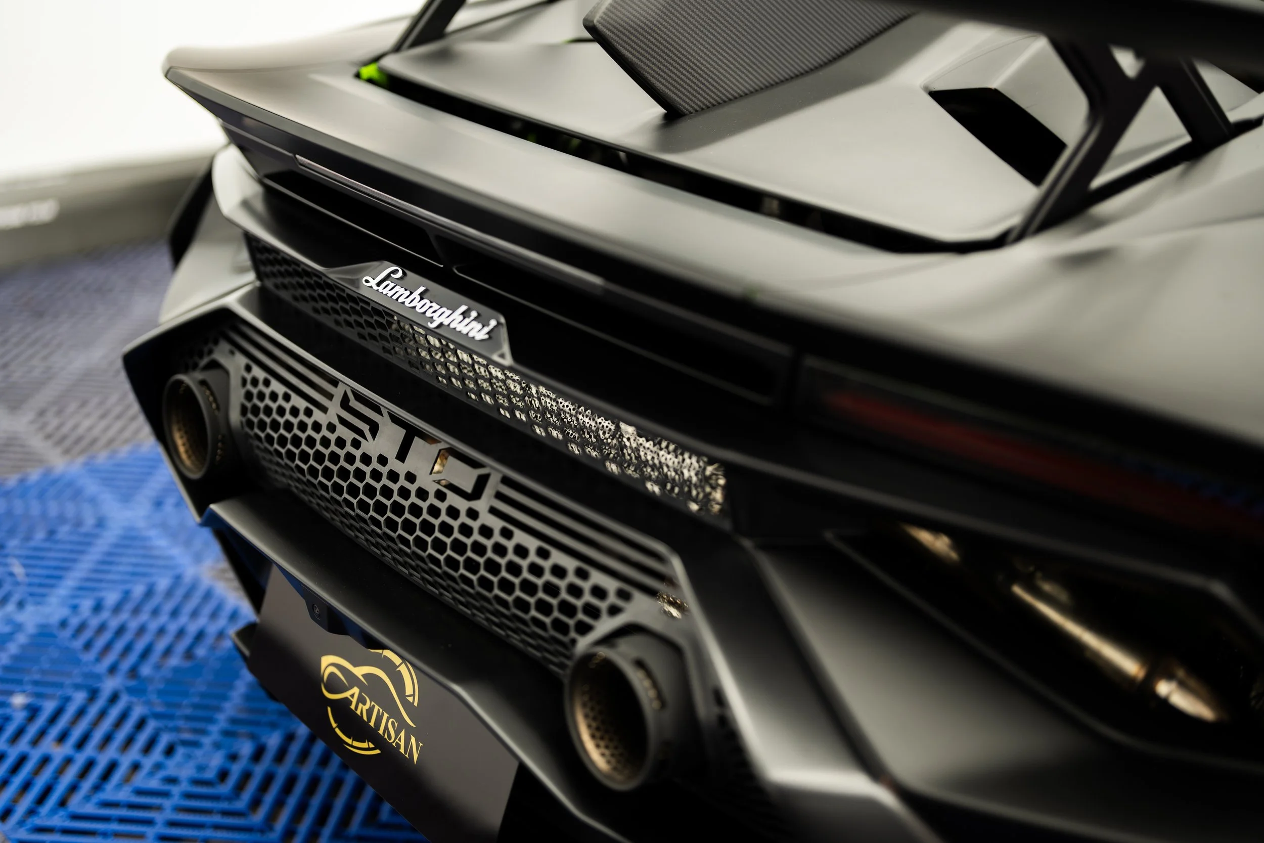 Close-up of the front of a black Lamborghini sports car with a prominent Lamborghini badge, custom grille, and dual exhaust pipes, on a blue and black checkered floor.