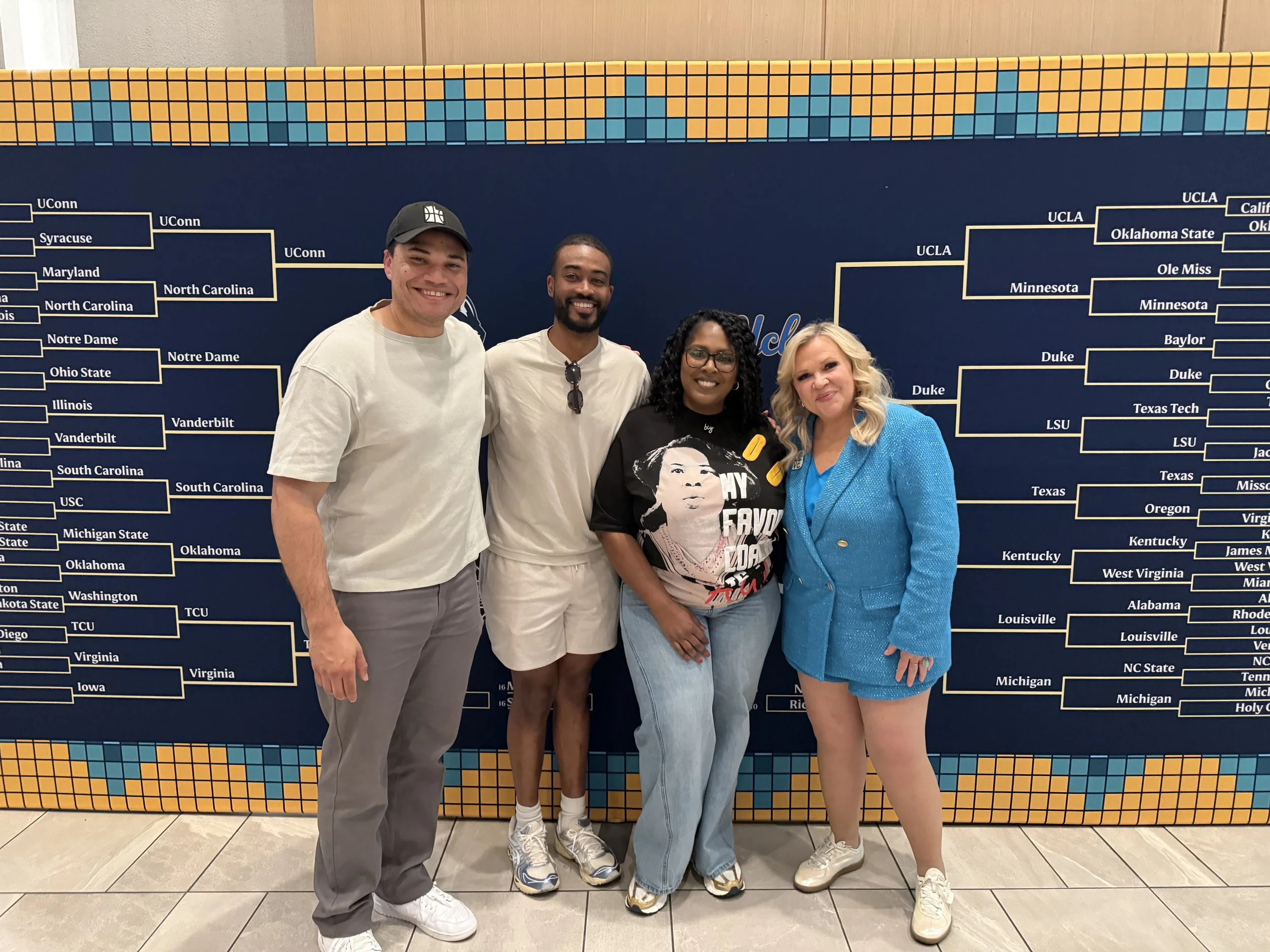 Four people standing in front of a large tournament bracket poster at an event. The bracket includes UConn, North Carolina, Notre Dame, Ohio State, Illinois, Vanderbilt, South Carolina, USC, Michigan State, Virginia, and other teams. The individuals are smiling and dressed casually.