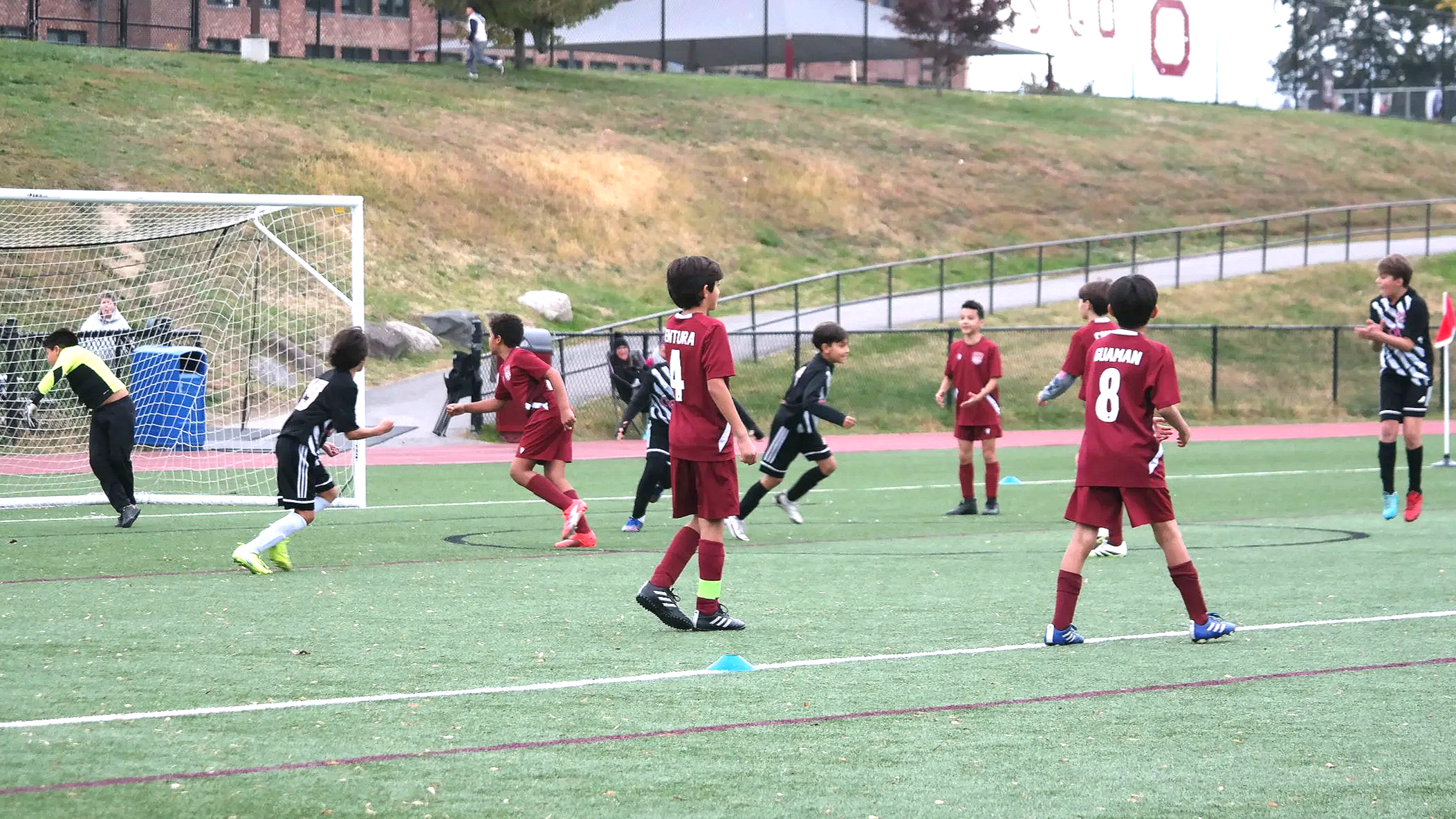Young boys playing soccer on a field during daytime. Some players are in black and white uniforms, while others are in red. A goalkeeper in bright yellow stands near the goal. The scene is outdoors with a grassy hill and a track in the background.