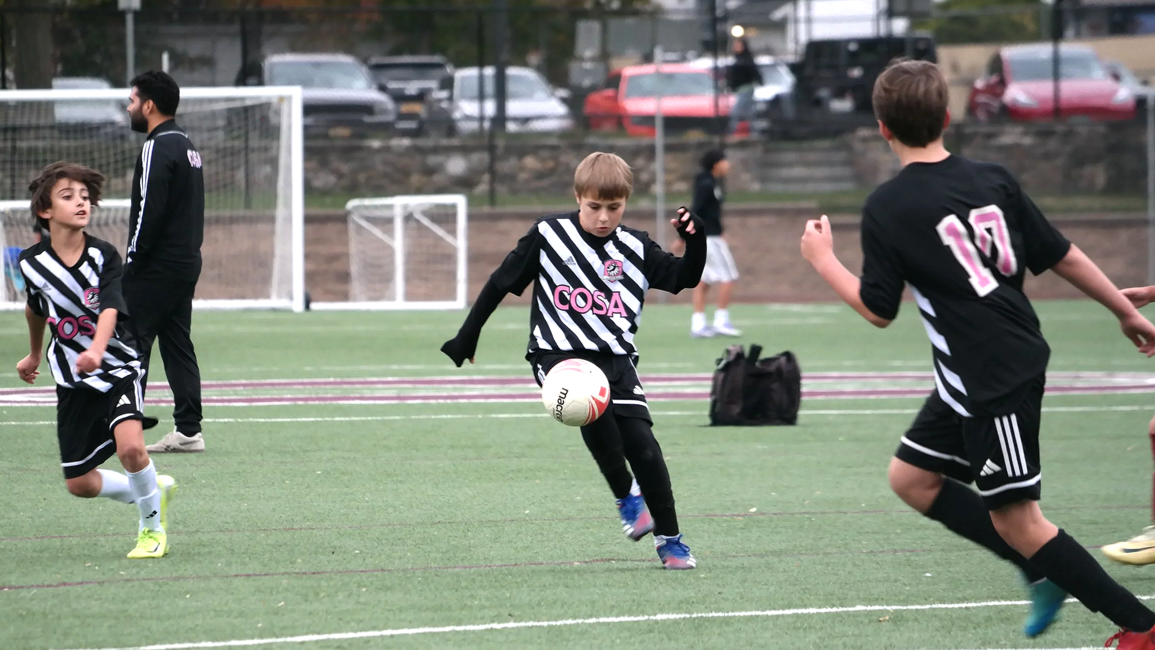 Young boys playing soccer on a field, wearing black and white striped jerseys with 'COSA' logo, with one boy kicking a ball while others run. A coach or adult stands nearby, and a few spectators are visible in the background.