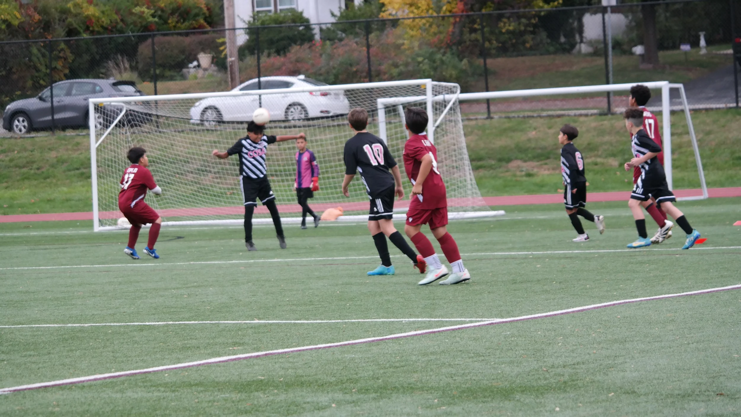 Children playing soccer on a field during daytime, with some kids in red jerseys and others in black uniforms, and a goalie in purple standing in front of the goal.