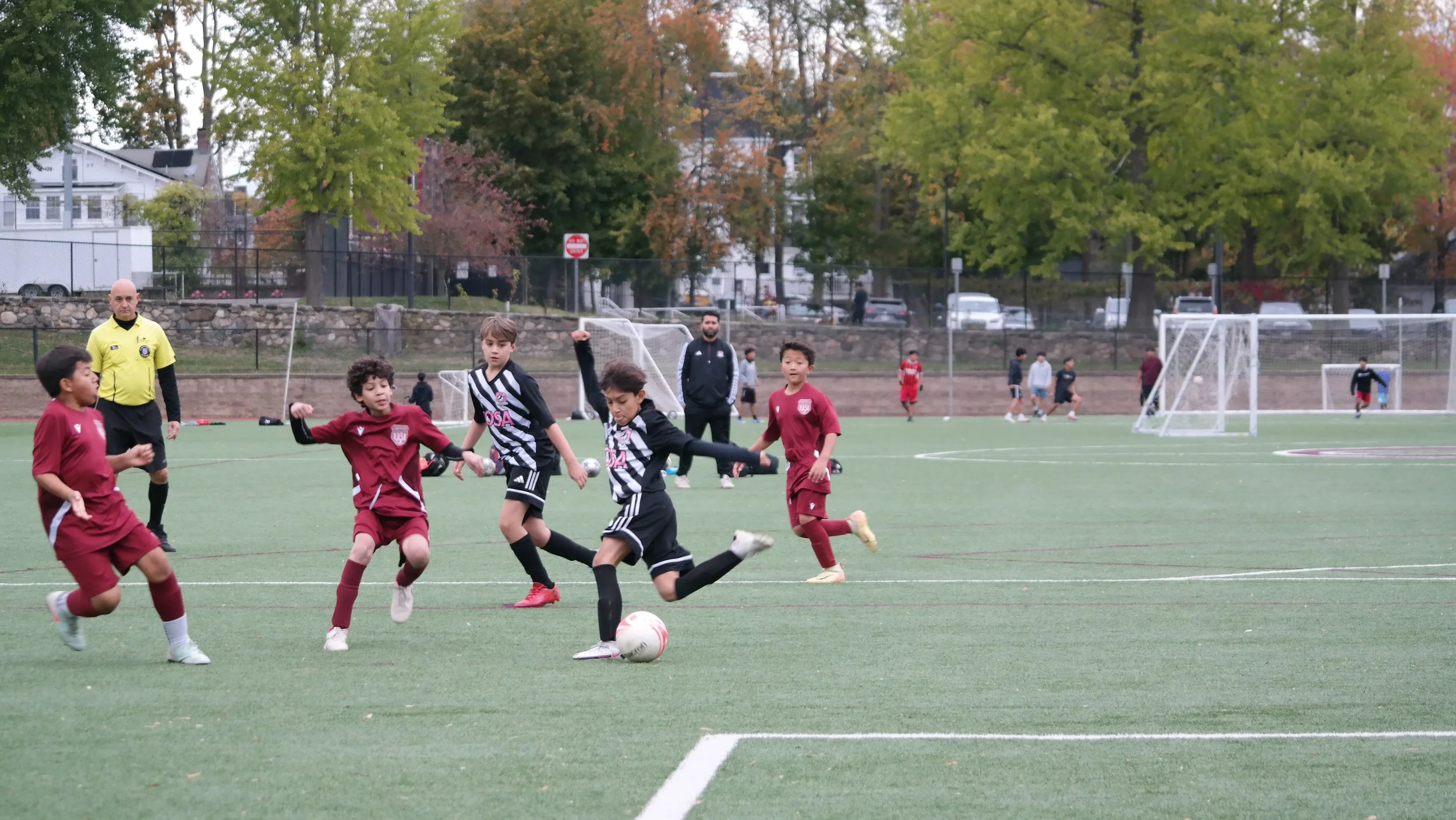 Young children playing soccer on a green field with goalposts in the background, with a referee and adults watching, surrounded by trees with fall foliage.