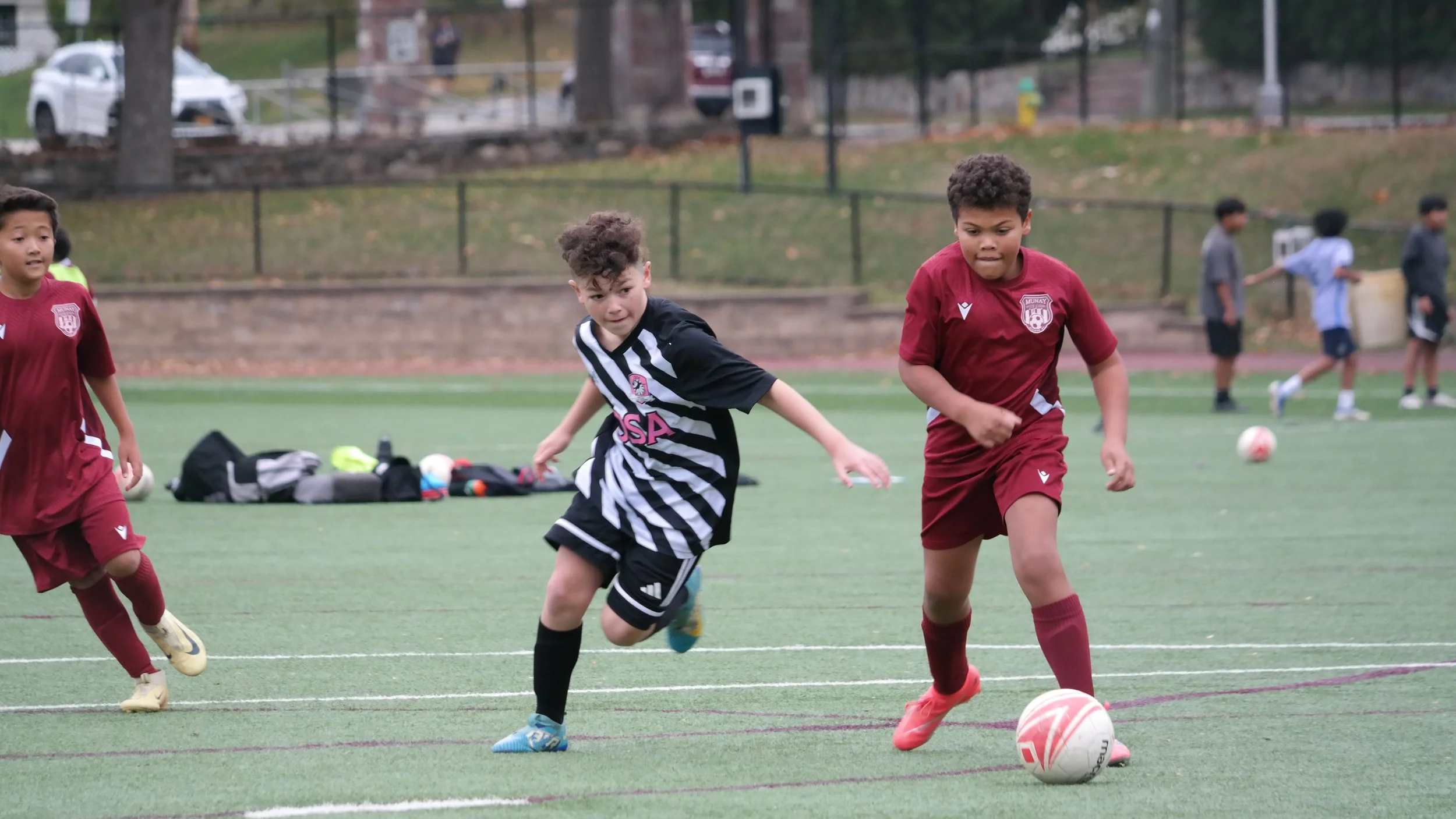 Young boys playing soccer on a green field, with one wearing a maroon uniform and dribbling a white ball with pink accents, and another in a black and white striped jersey reaching for the ball.