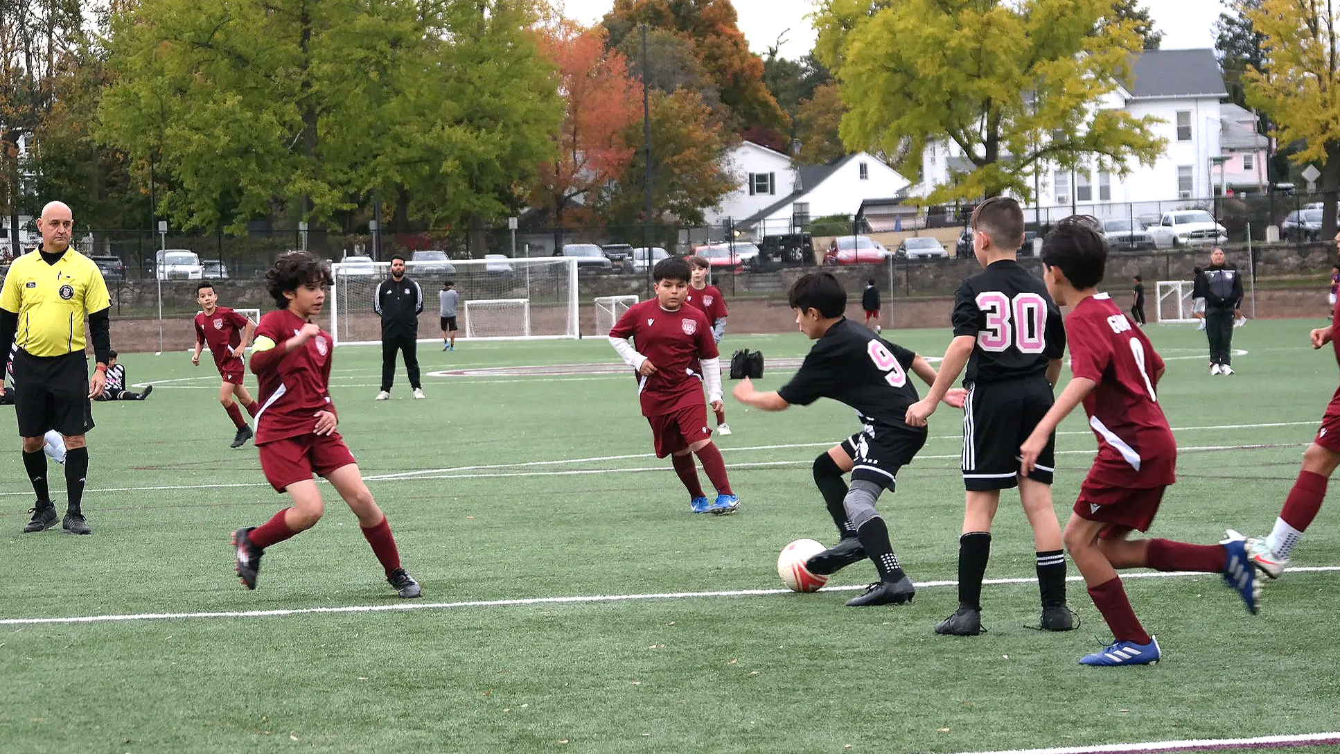 Children playing soccer on a field with a referee watching. Kids are in maroon and black uniforms, competing for the ball. Trees with fall foliage and houses are in the background.