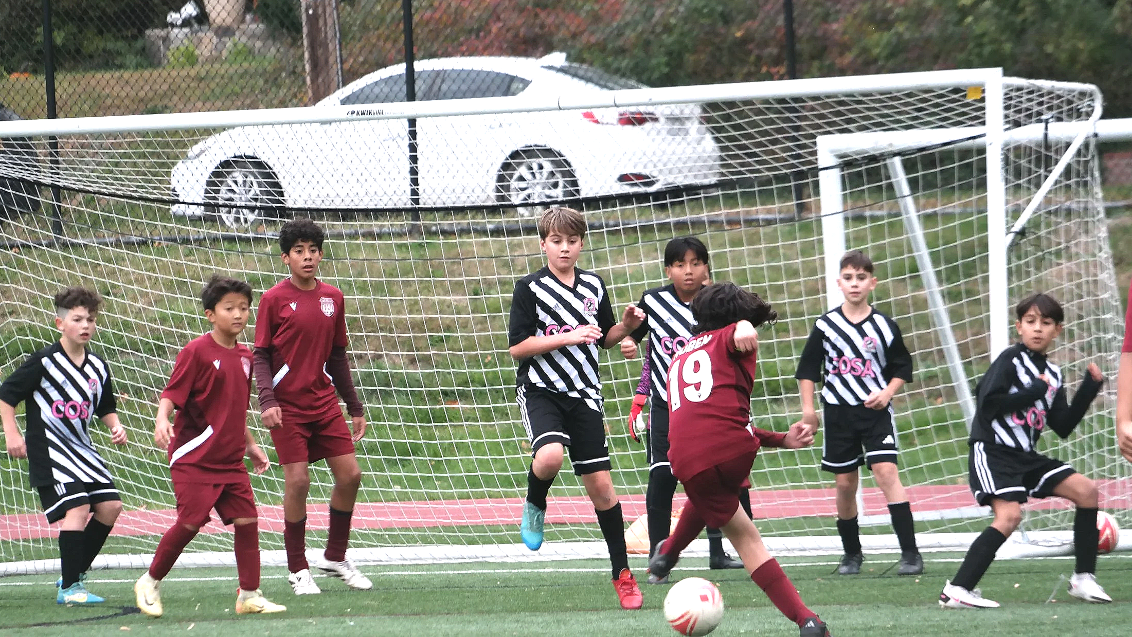 Children playing soccer on a field, with some trying to score a goal and others guarding the net, during daytime.
