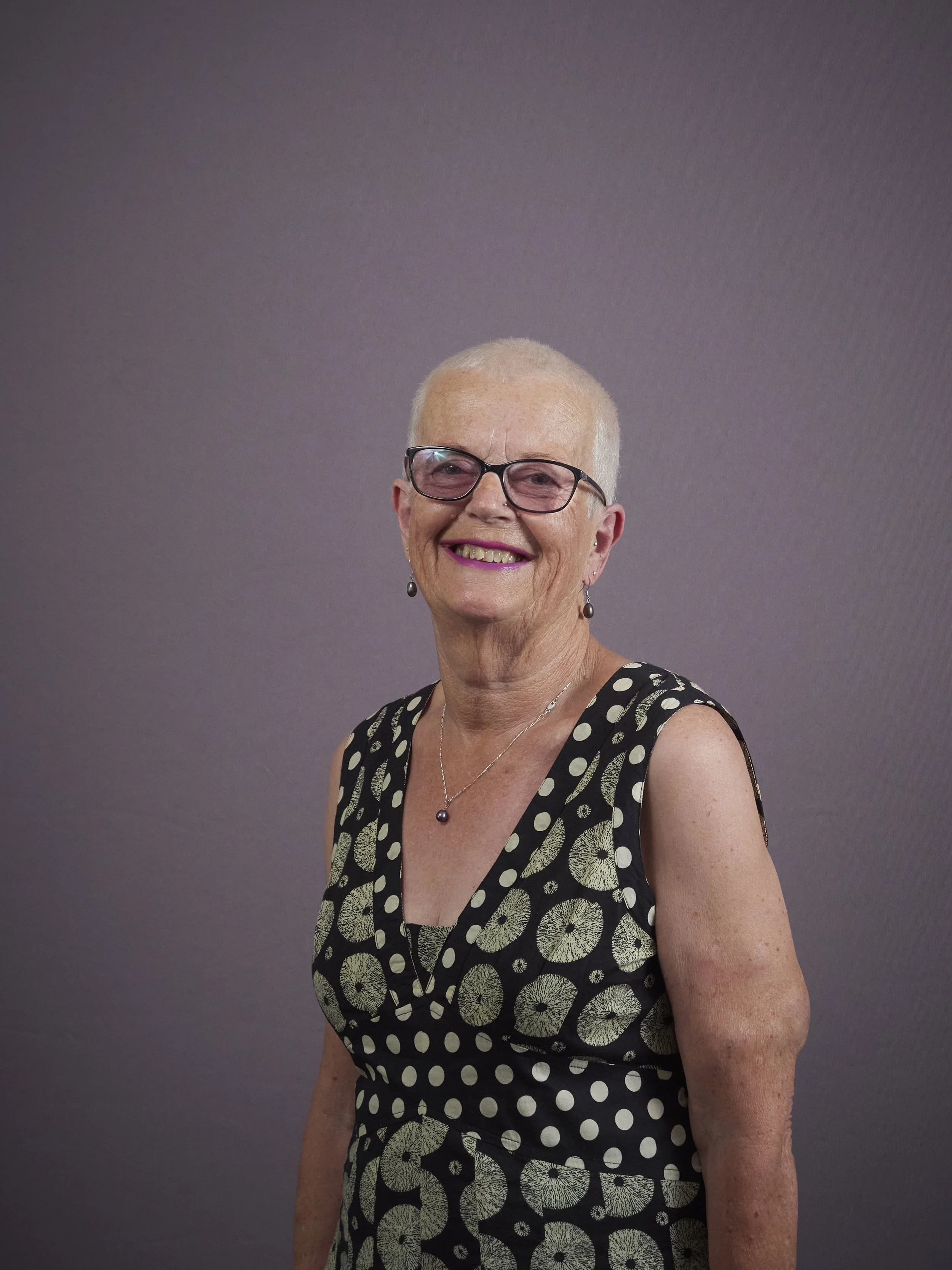 Smiling elderly woman with short hair wearing glasses, a light blue sweater, pendant necklace, and earrings, against a blue background.