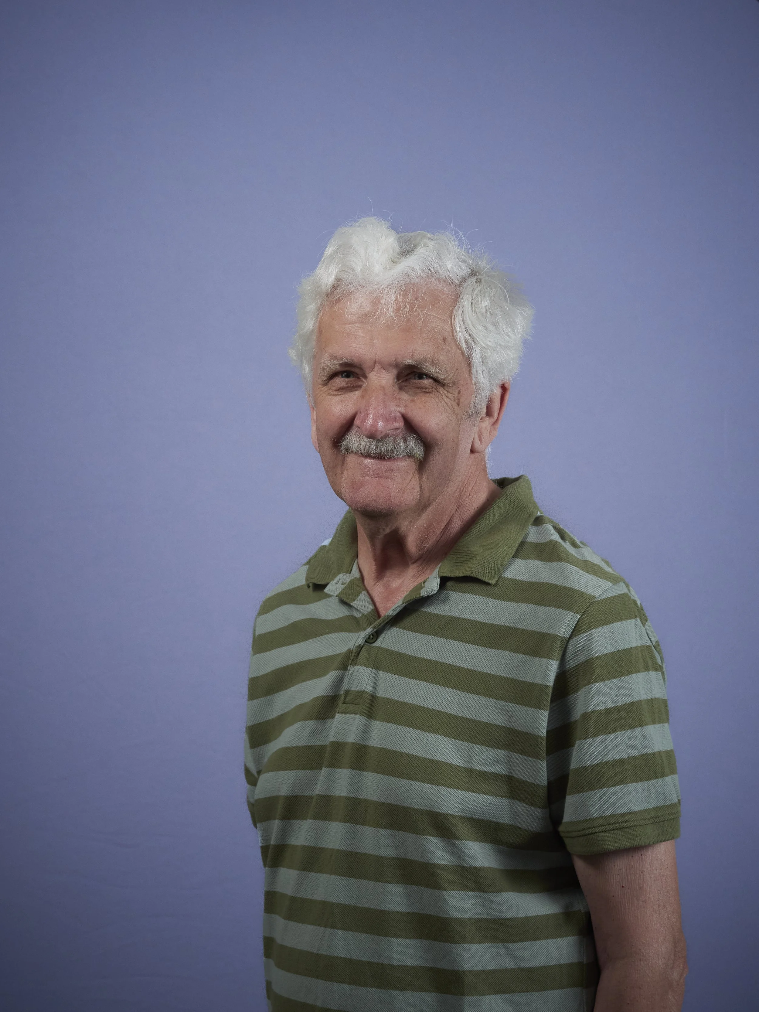 Portrait of an elderly man with white hair and a mustache, wearing a blue zip-up jacket and a black shirt, against a plain blue background.