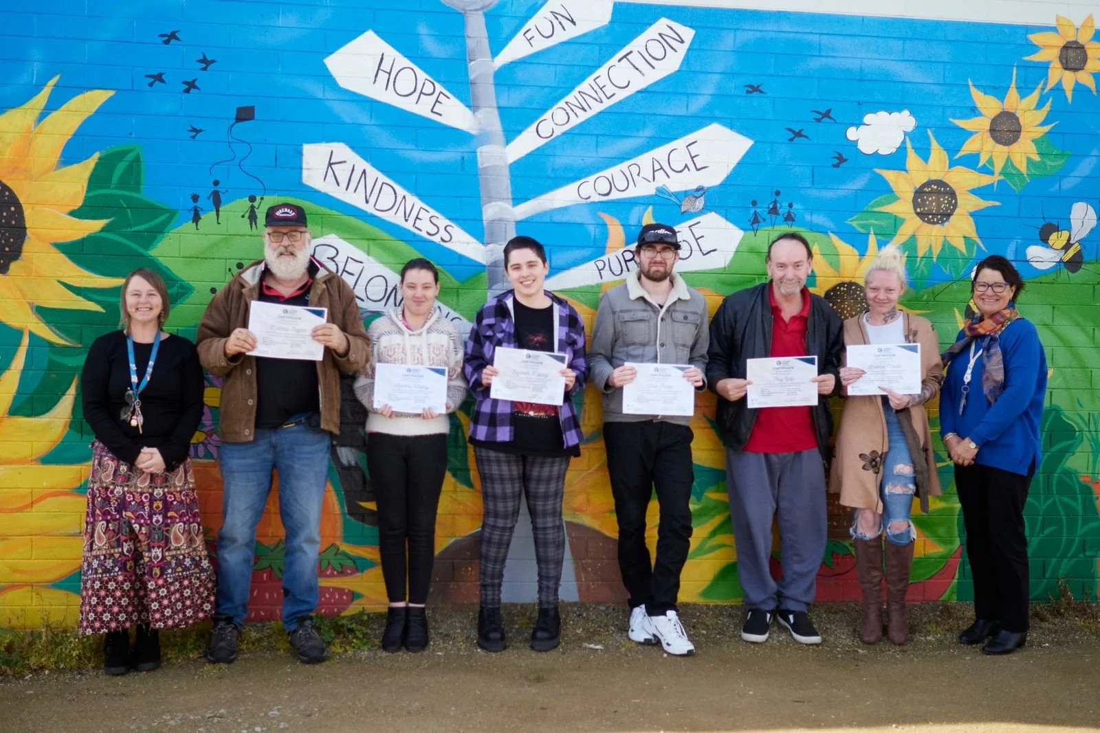 Group of five people standing in front of a colorful mural with a tree and sunflower theme. The mural has signs with words like hope, kindness, fun, connection, courage, and purpose. The three women in the middle are holding certificates.