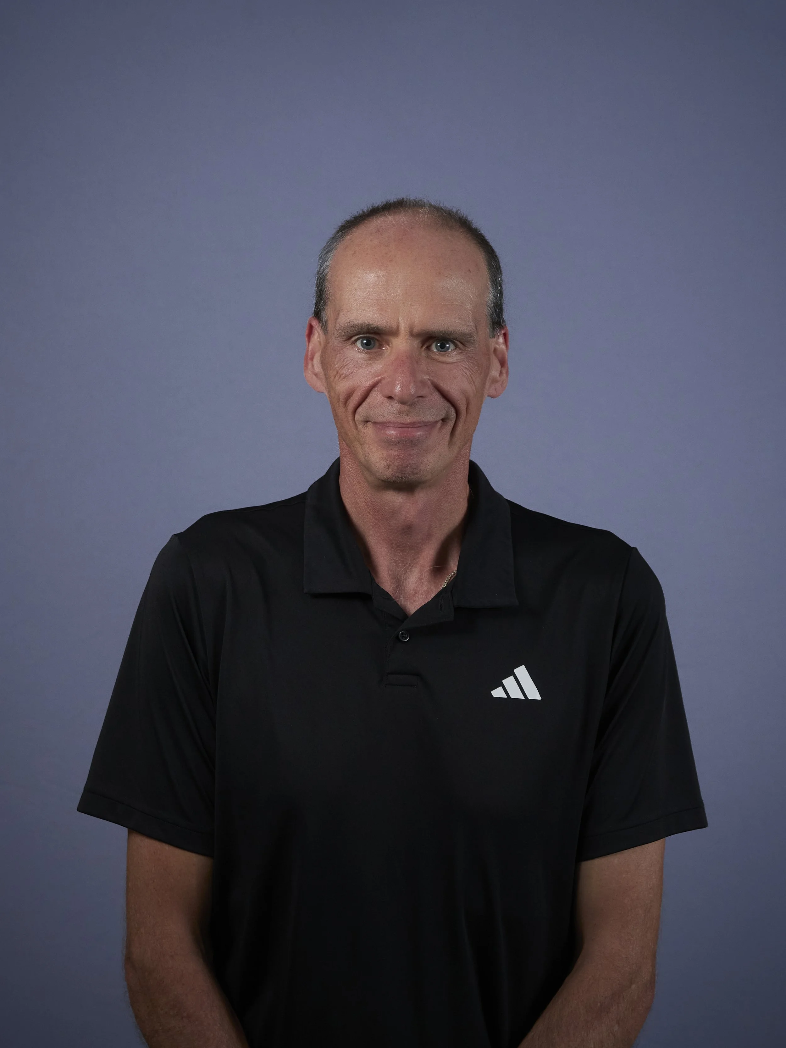 Portrait of a man wearing a black City Mission cap, smiling outdoors with cloudy skies and trees in the background.