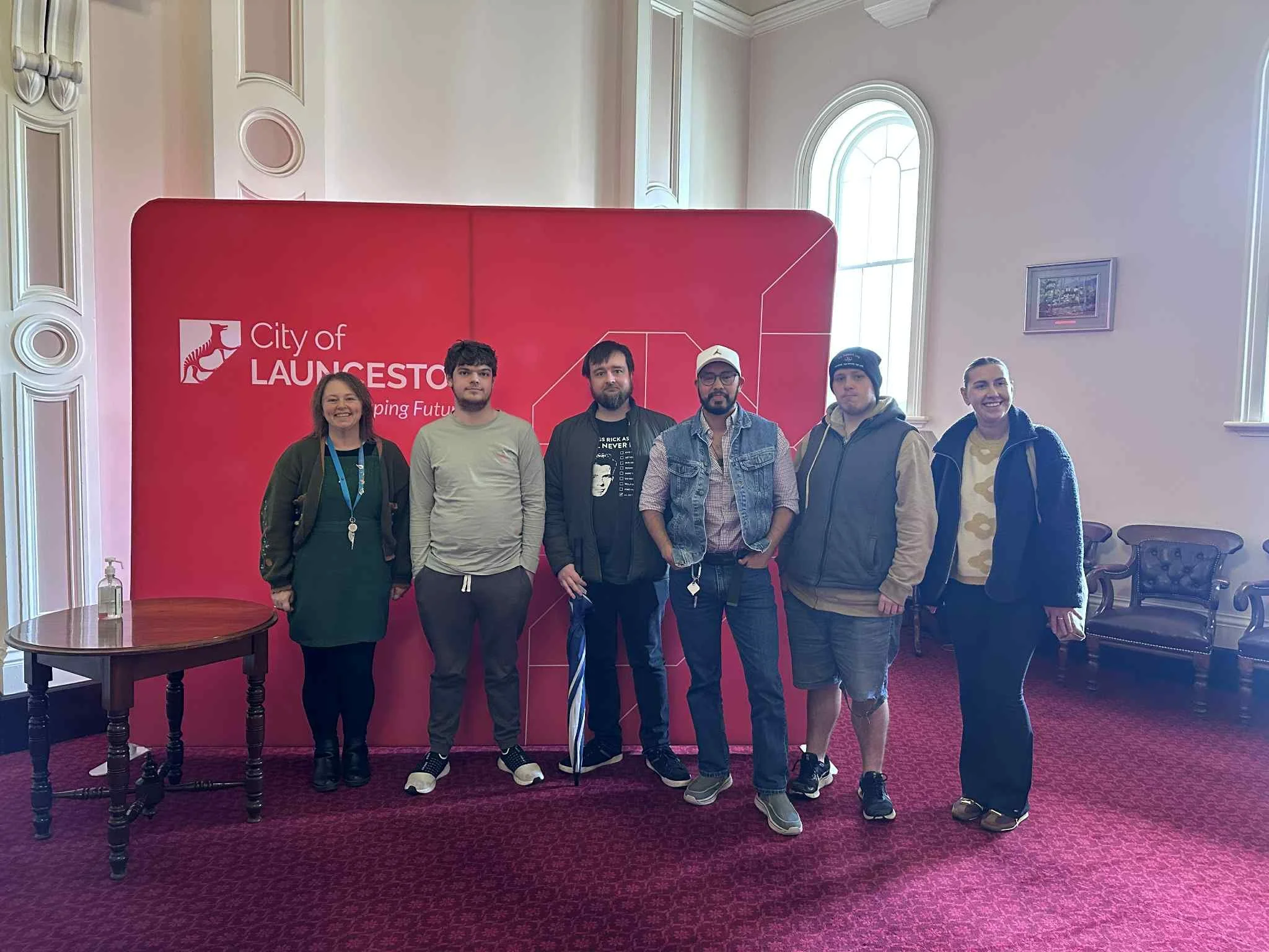 Group of eight women standing outdoors on a sunny day, with a brick building, trees, and a sidewalk in the background.