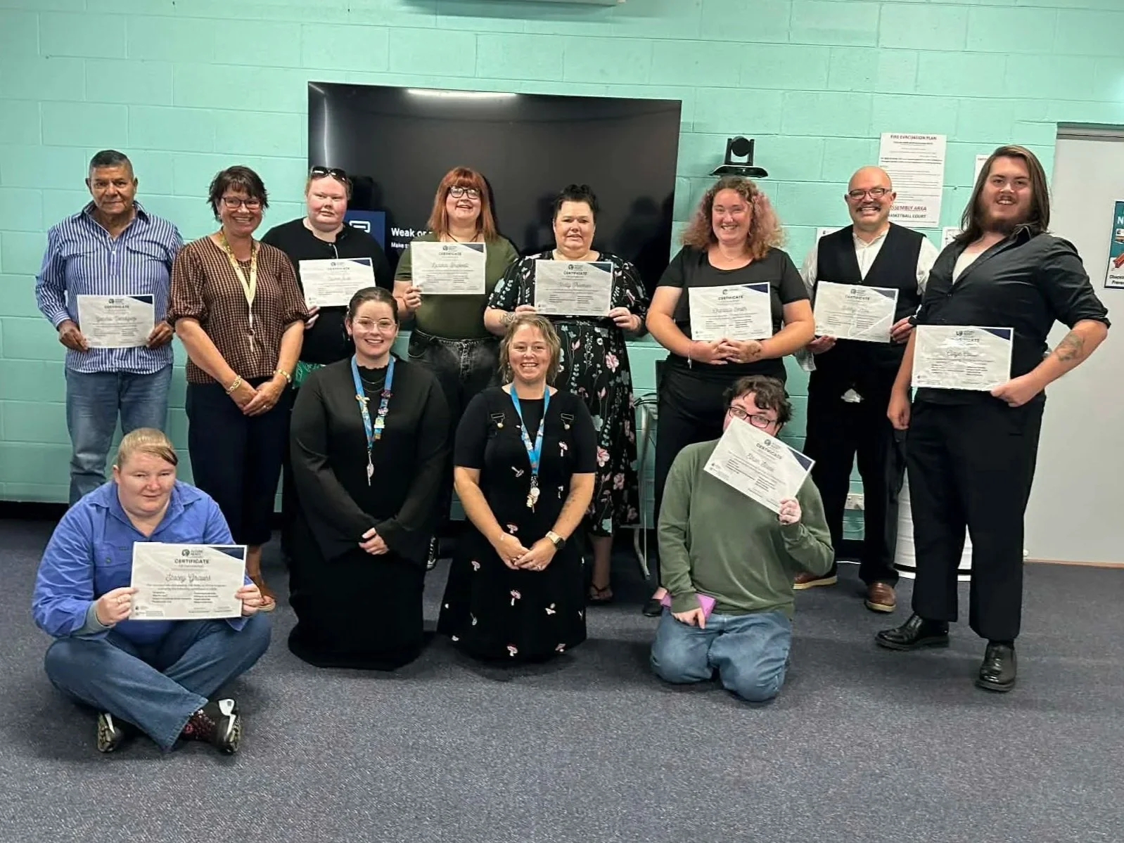 Group of five people standing in front of a colorful mural with a tree and sunflower theme. The mural has signs with words like hope, kindness, fun, connection, courage, and purpose. The three women in the middle are holding certificates.
