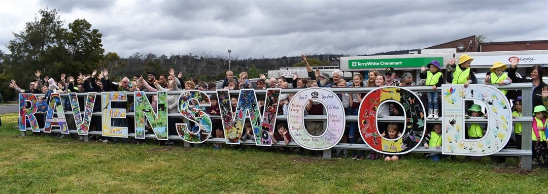 A large group of people stand outdoors on a grassy area behind a colourful “RAVENSWOOD” sign. Each letter is decorated with different patterns and drawings, suggesting a community art project. Trees and buildings are visible in the background.