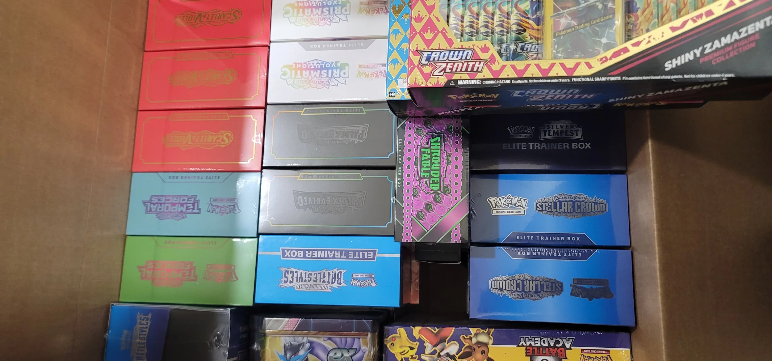 Assorted Pokémon trading card game boxes stacked on a table, including Crown Zenith, Silver Tempest, Scarlet Violet, Battle Academy, and others.