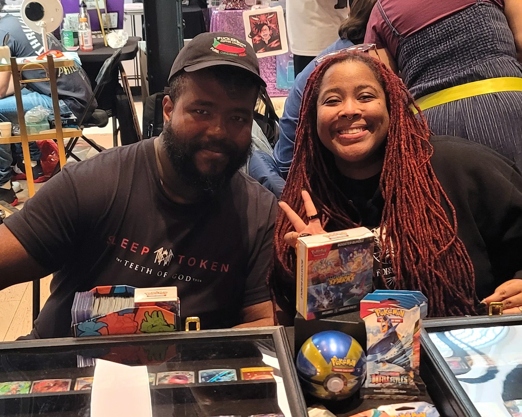 A man and a woman sit at a table with Pokémon cards, booster packs, and merchandise. The woman has long red braided hair, is smiling, and making a peace sign. The man has a beard, is wearing a black cap, and is looking at the camera. They are at a Pokémon trading card event.