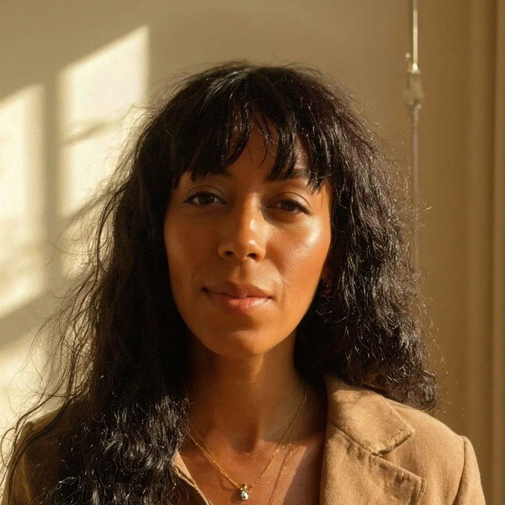 Image of Sana Keefer with long, dark, curly hair smiling softly in natural sunlight, wearing a beige jacket and layered gold necklaces. This is a photo of Sana Keefer, creative and brand executive.