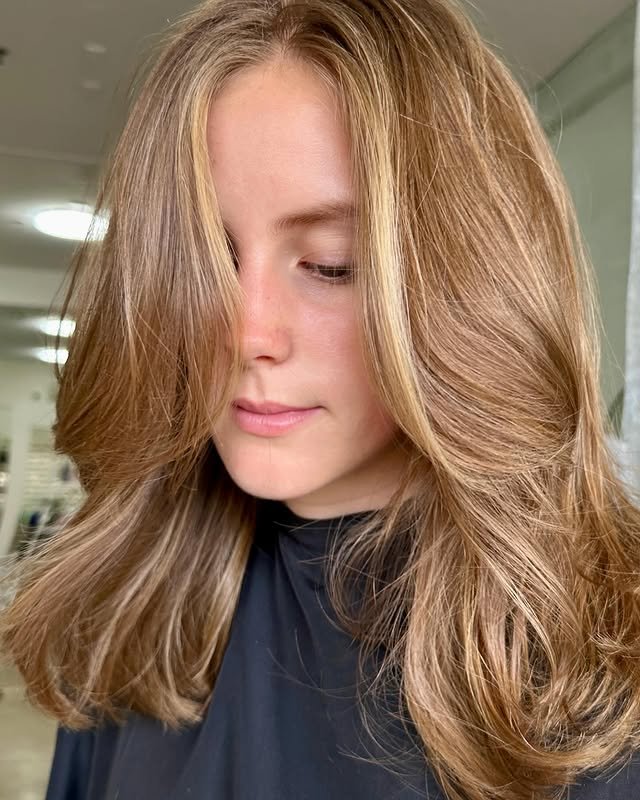 Woman with light brown wavy hair styled in a layered haircut, wearing a black cape in a salon setting.