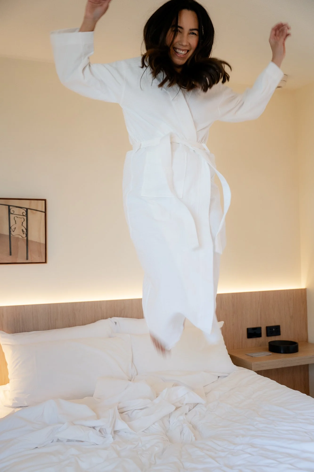 A woman in a white bathrobe jumping on a bed, smiling and enjoying herself in a hotel room.