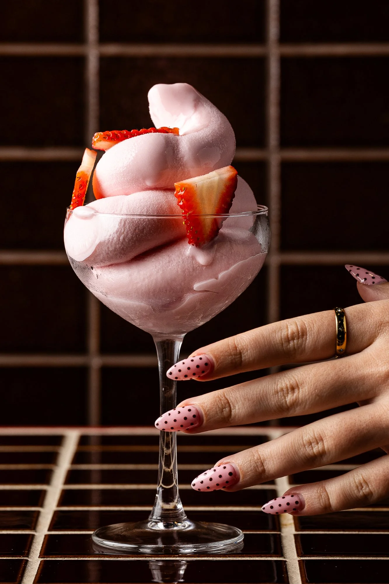 A hand with pink polka dot nails and a gold ring holds a wine glass filled with pink strawberry-flavored frozen dessert topped with sliced strawberries, set against a dark background with a wooden grid.