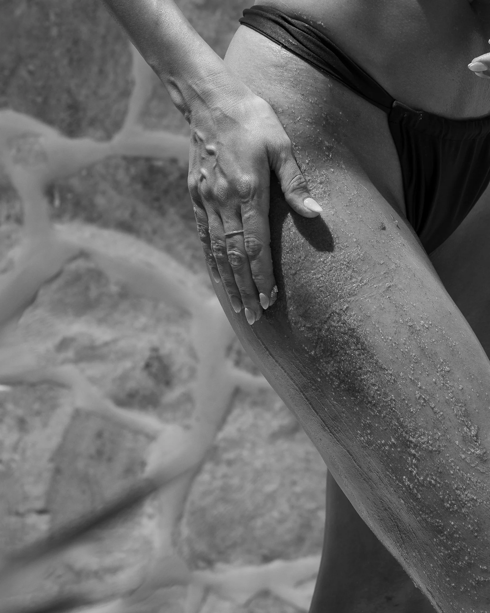 Close-up of a person's hand applying lotion or sunscreen on their thigh, with a stone wall in the background.