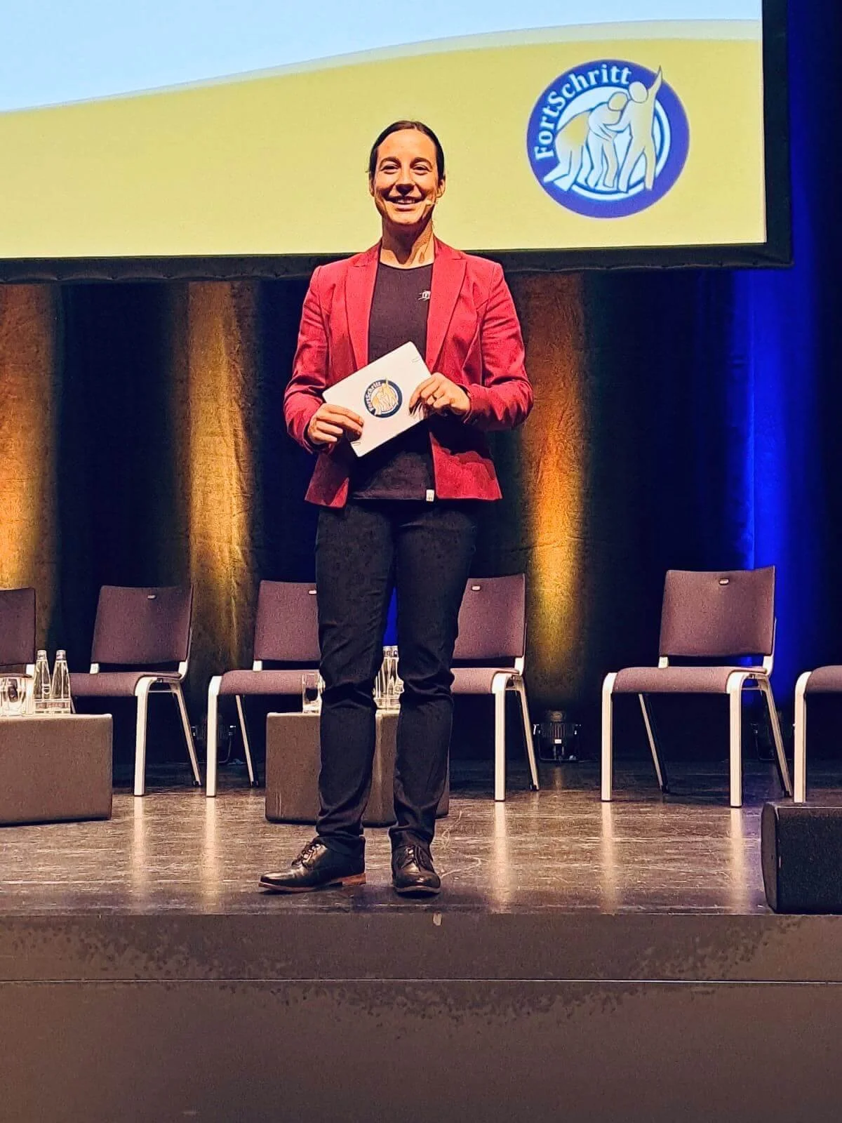 Moderatorin Janine Faaß steht auf einer Bühne in deren Hintergrund man das Logo von Fortschritt Bayern sieht.
