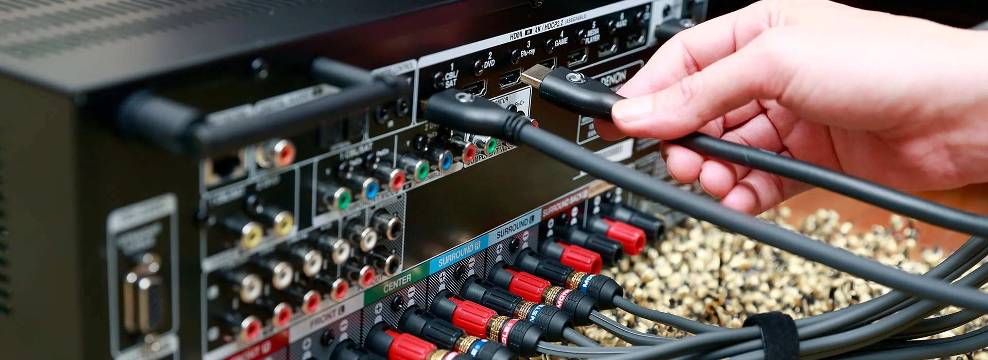 A person's hand plugging a black cable into the back of an audio/video receiver with multiple colorful cables and ports.