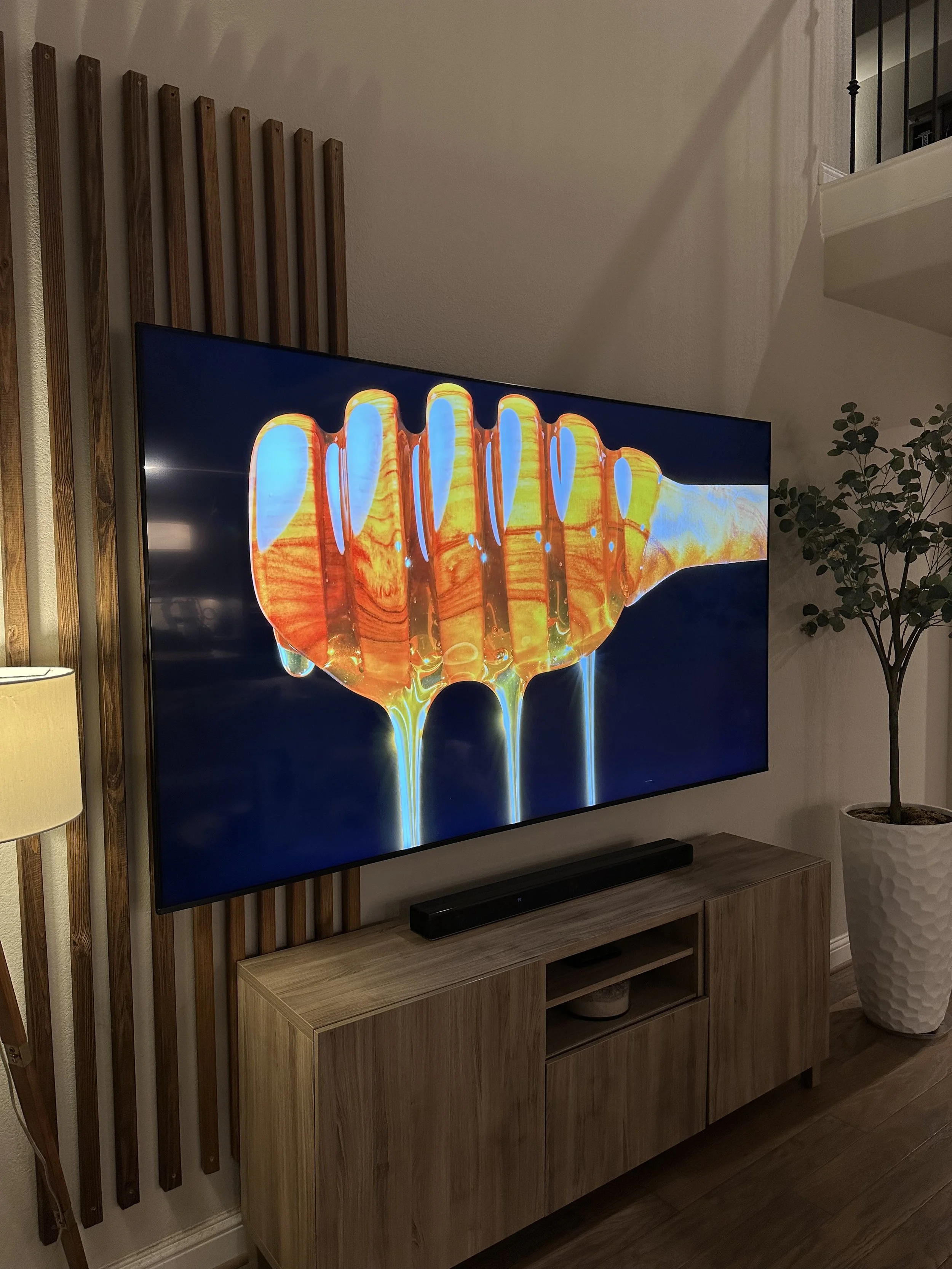 Modern living room with a wall-mounted flat-screen TV displaying vibrant orange and yellow honey dripping from a honey dipper, a wooden media console below, a soundbar, a tall potted plant, and vertical wooden wall accents.