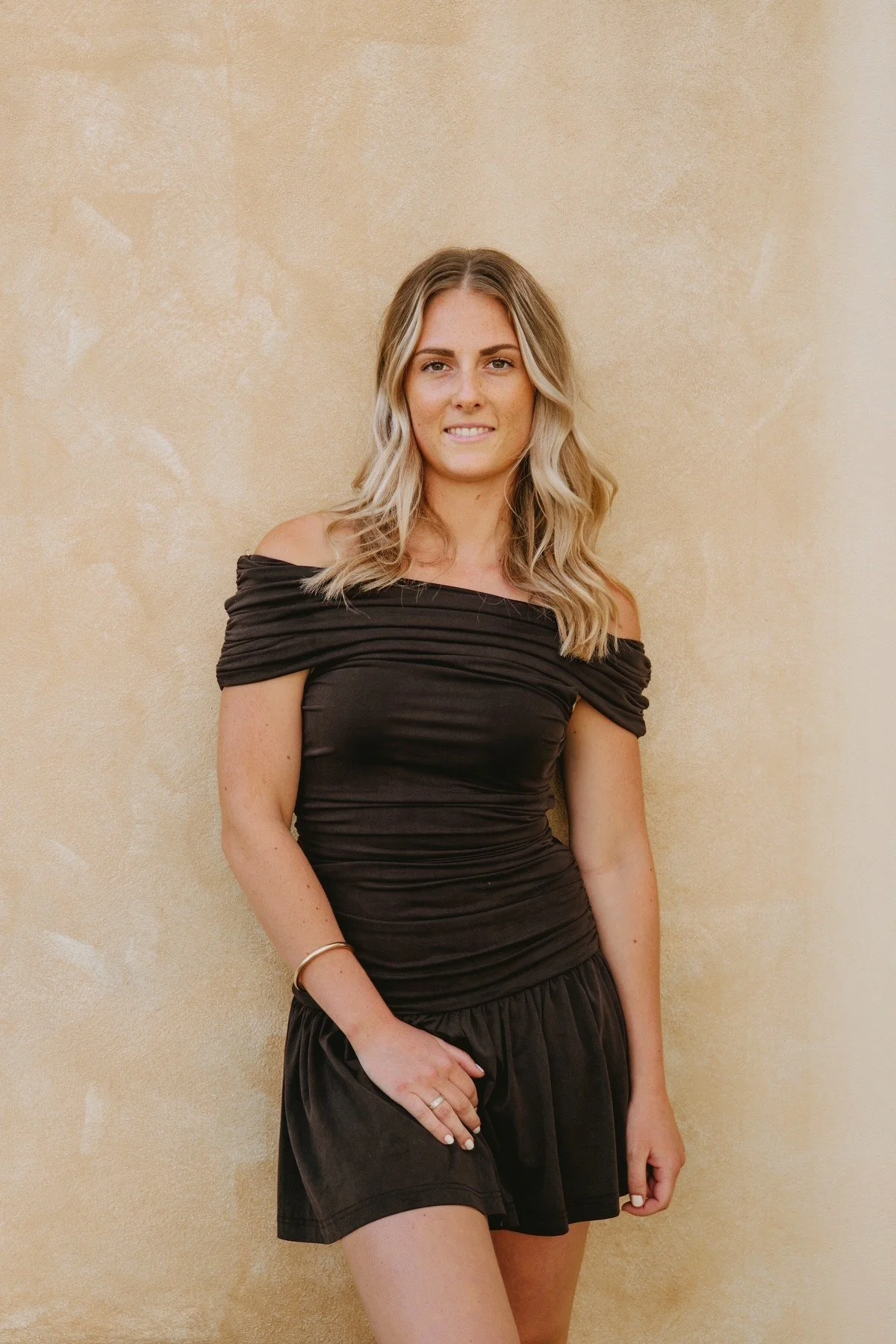 A woman with blonde hair styled in loose waves, wearing a black off-shoulder dress with ruched fabric, standing against a beige textured wall.