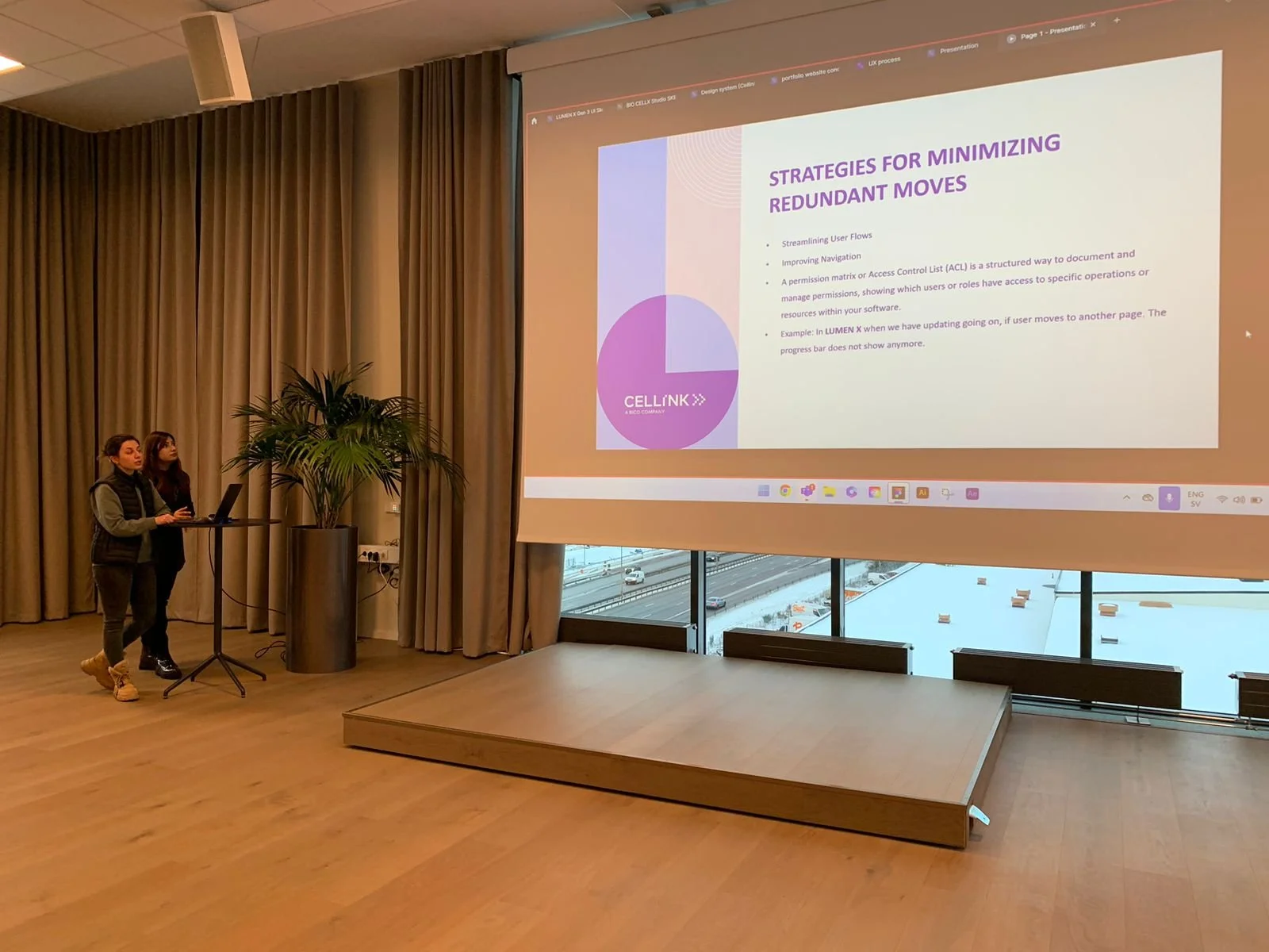 Two women standing near a presentation screen, one is using a laptop, with a large window and a snowy outdoor scene in the background. The presentation slide displays strategies for minimizing redundant moves.
