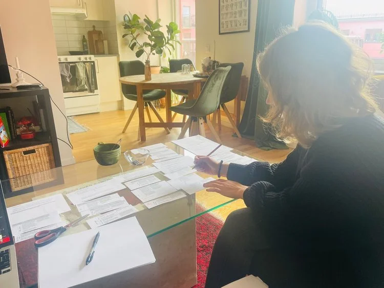 A woman with curly blonde hair working at a glass-top table covered with papers and documents in a cozy living room with a dining area, a plant, and a window letting in natural light.