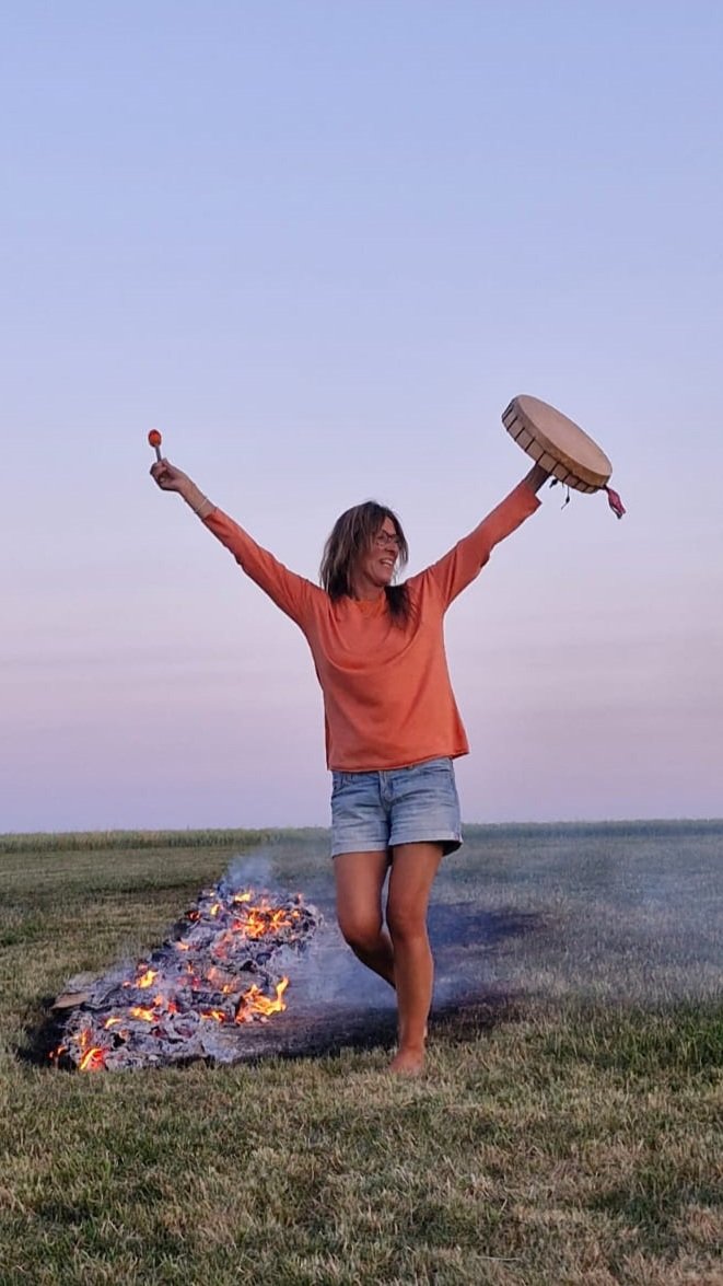Eine lachende Frau in einem weißen Kleid und einer orangefarbenen Bluse tanzt barfuß auf einer Wiese neben einem Lagerfeuer, das in der Nähe brennt und den Himmel im Hintergrund erleuchtet.
