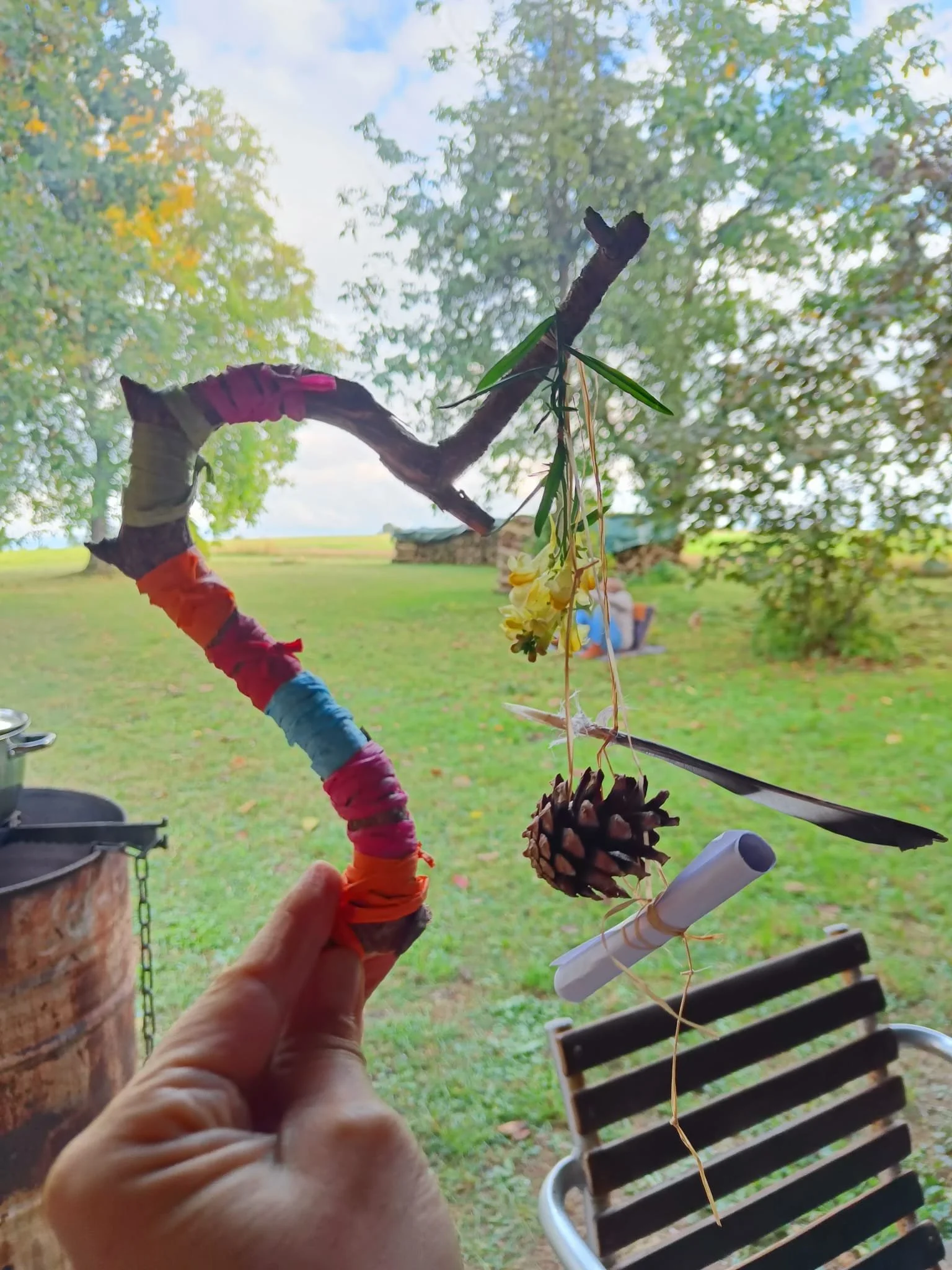 Eine handgefertigte Dekoration besteht aus einem verzweigten Ast, der bunt eingewickelt ist, mit einer Kiefernzapfen und einem Papierrohr, die an einer Schnur hängen. Im Hintergrund ist eine grüne Wiese mit Bäumen und einem Himmel mit Wolken zu sehen