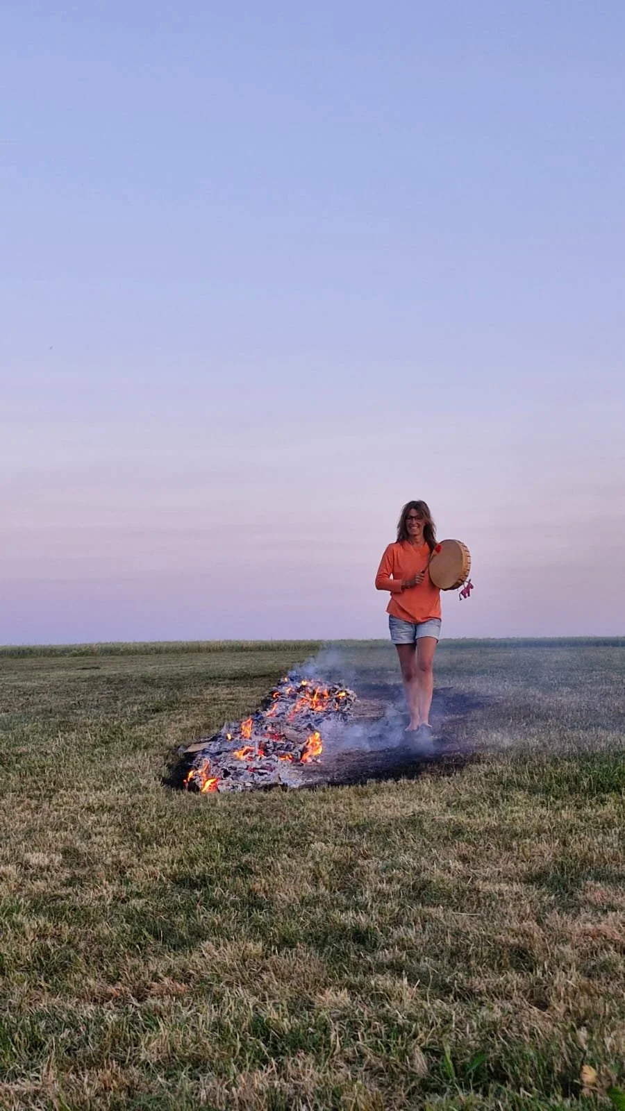 Eine lachende Frau in einem roten Shirt und Jeans-Shorts läuft barfuß an einem brennenden Holzstoß auf einer Wiese bei Sonnenuntergang, während sie eine Trommel hält.