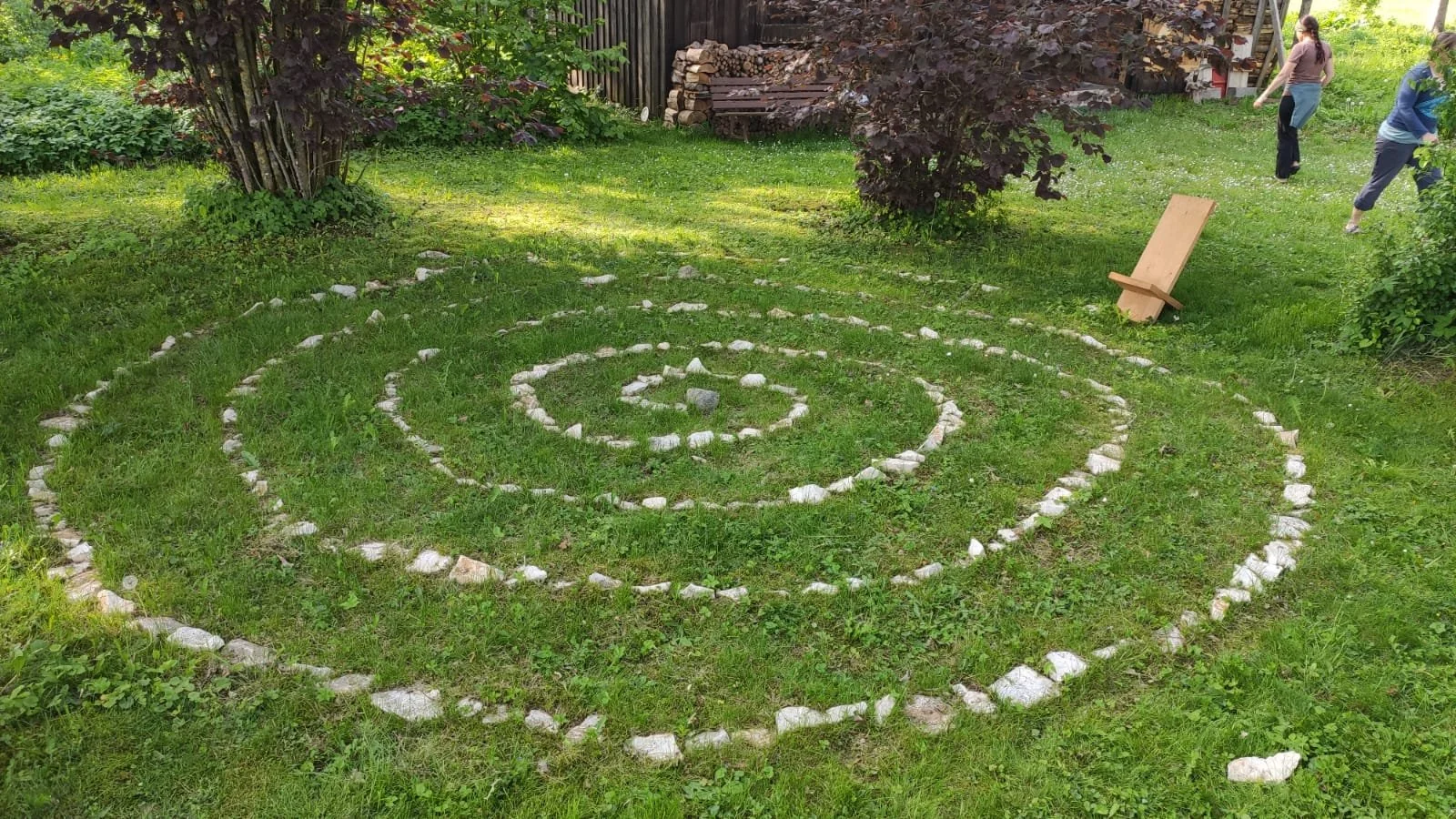 Ein grüner Garten mit einem Spiralförmigen Muster aus weißen Steinen auf dem Rasen, im Hintergrund einige Bäume, eine Frau und ein Mädchen, die spielen, sowie ein Holzstuhl, der auf dem Rasen steht.