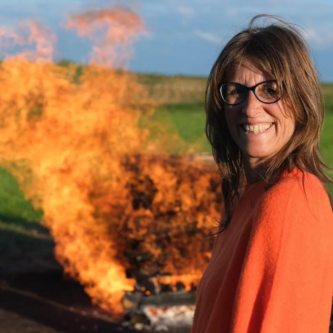 Eine lachende Frau mit Brille und braunem Haar trägt einen orangefarbenen Pullover, im Hintergrund ein brennender Zug auf freiem Feld bei Sonnenuntergang.