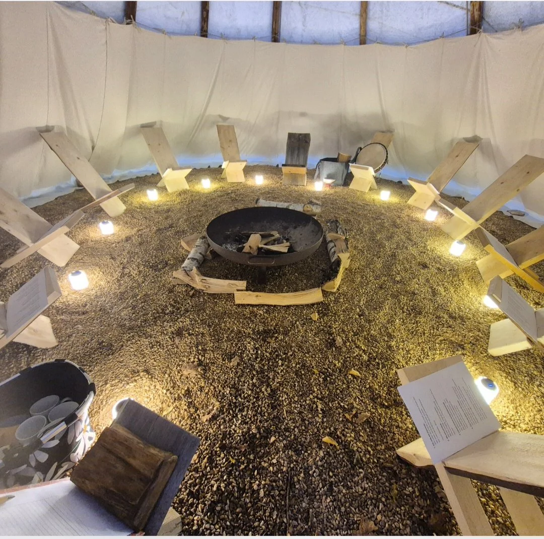 Innenansicht eines zeltartigen Raums mit einem Lagerfeuer in der Mitte, umgeben von Stühlen aus Holz und Kerzenlichtern, mit Notizen oder Broschüren auf den Stühlen und einem weißen Stoff als Wandverkleidung.