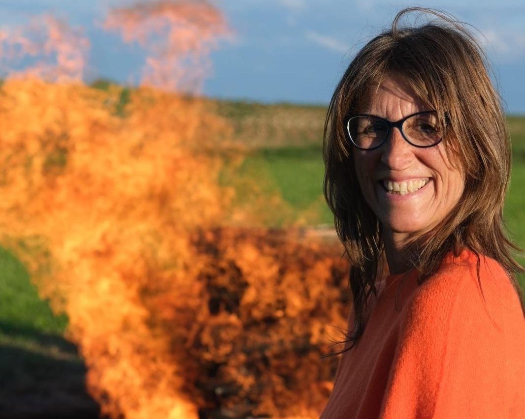 Eine lachende Frau mit Brille vor einem brennenden Feuer, im Hintergrund eine grüne Landschaft und einen blauen Himmel.