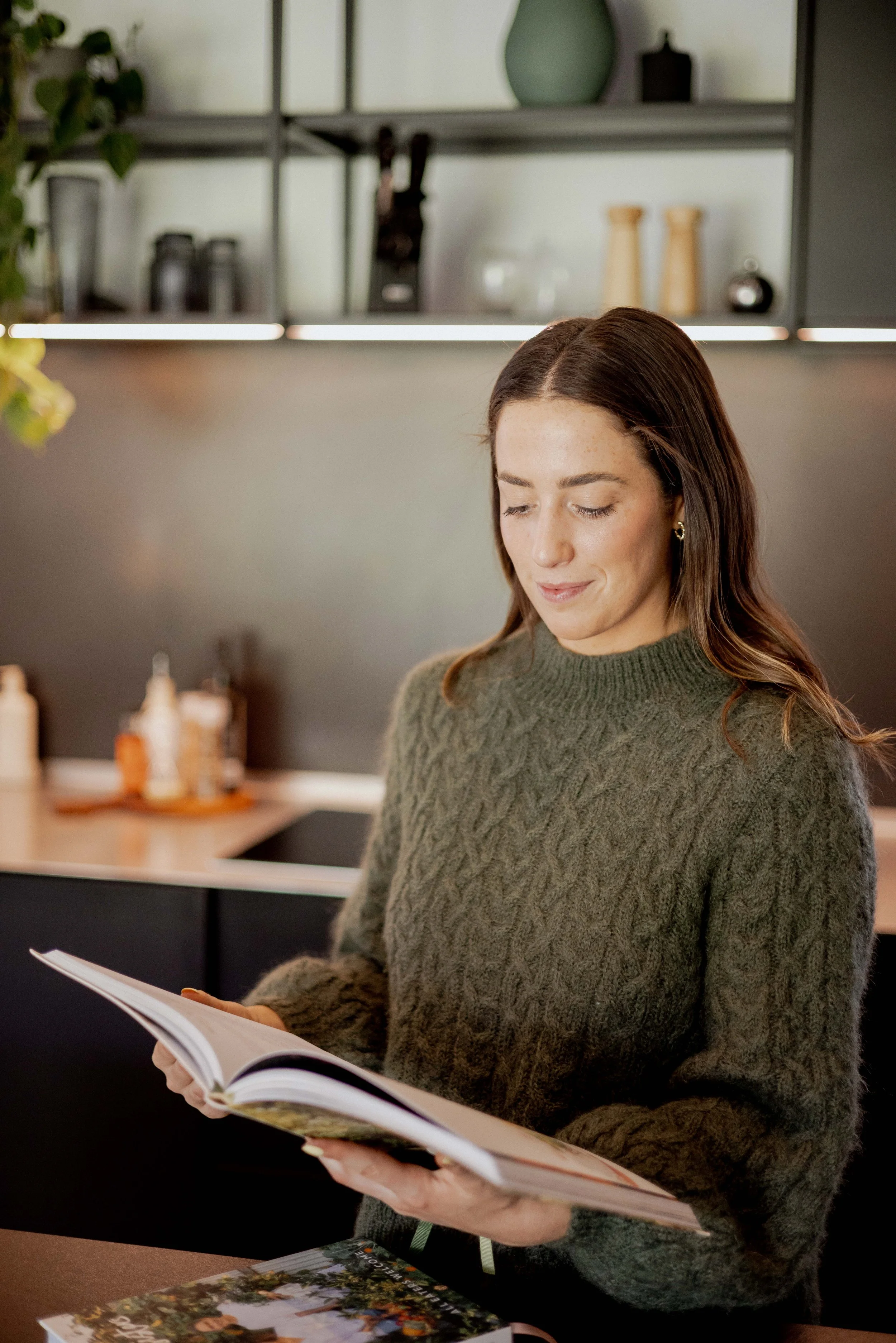 Woman reading a book in a cozy kitchen with dark cabinets, decorative bottles, and green plants.