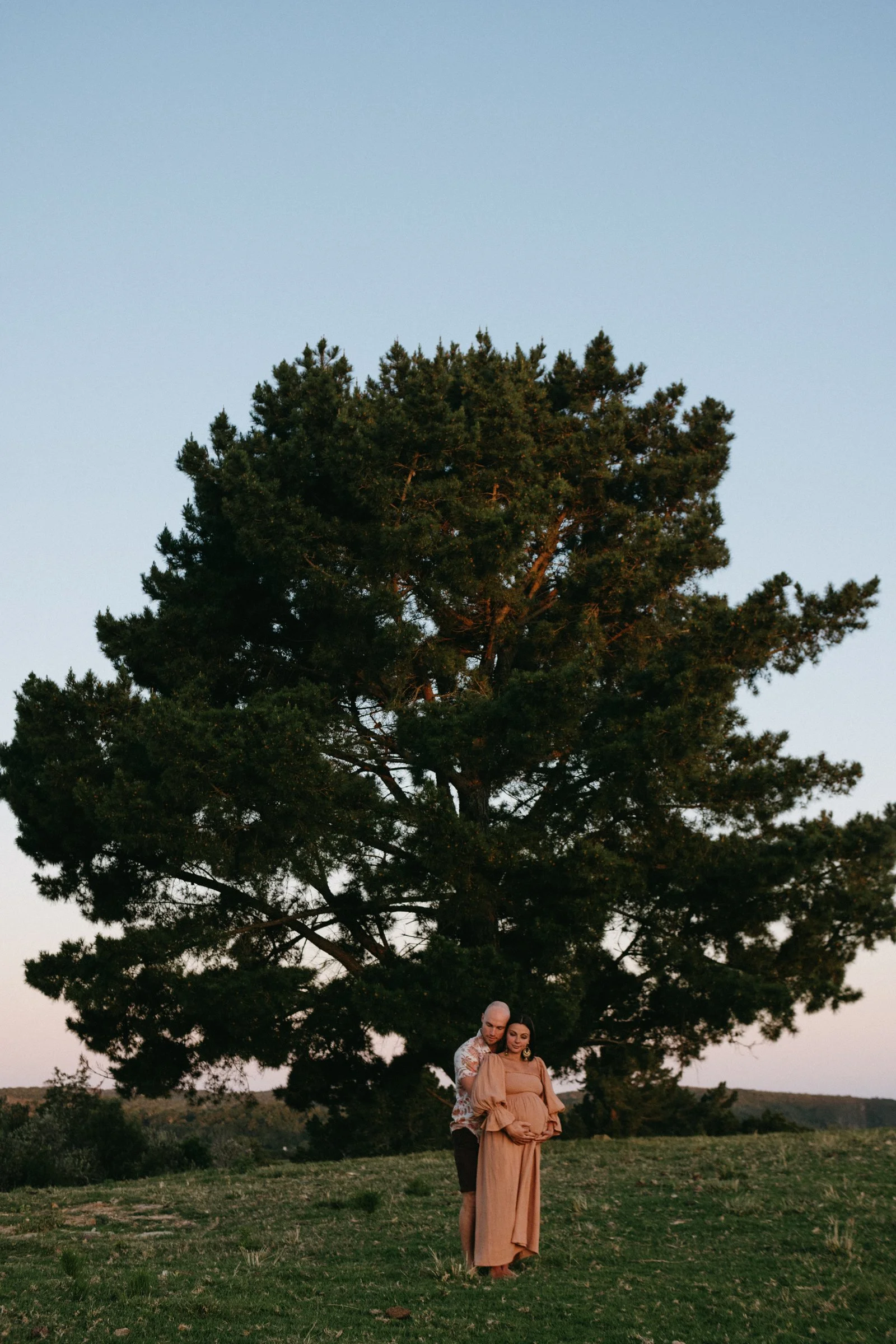 Pregnant woman, with her husband standing behind her in front of a tree.