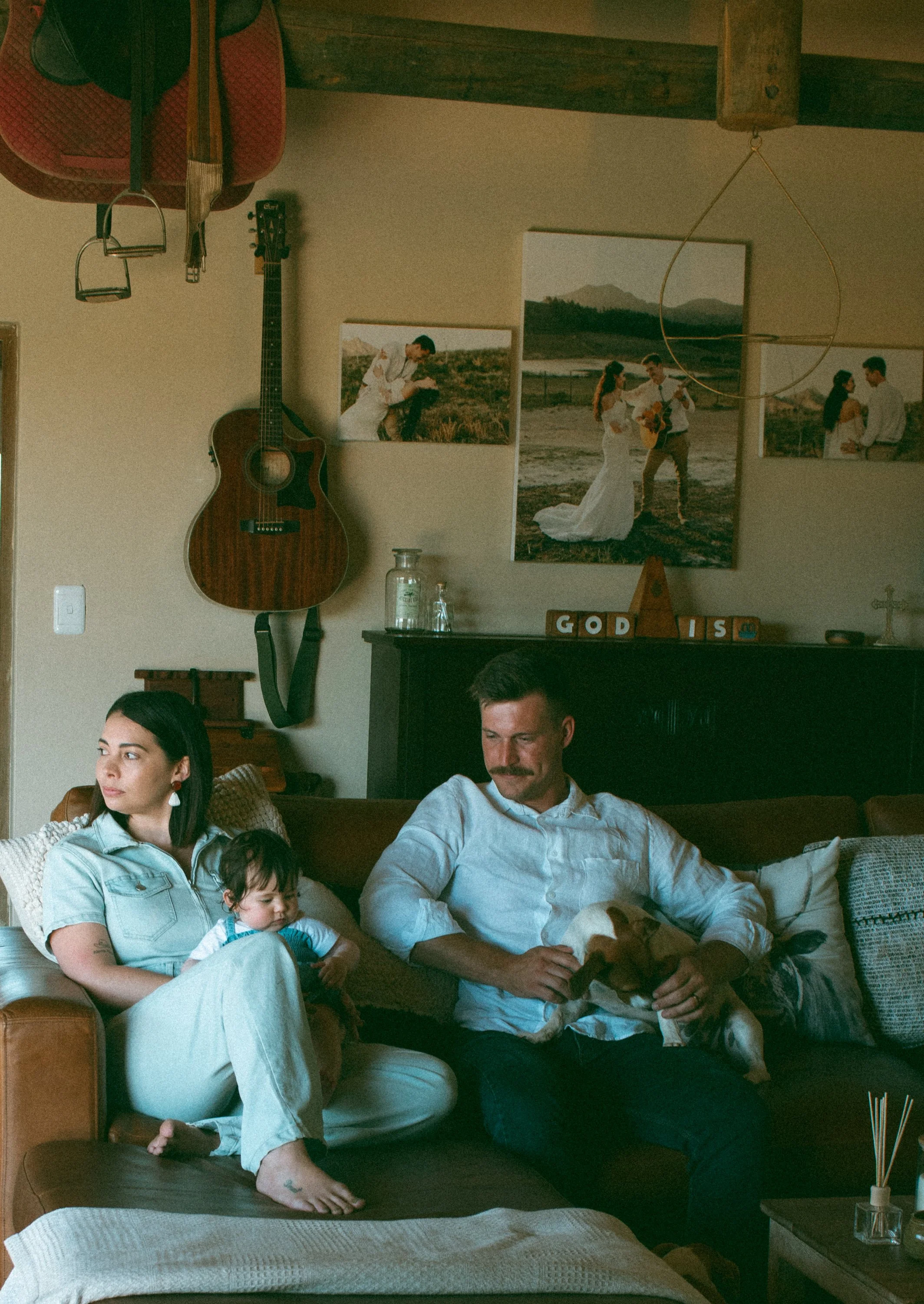 A family of three sitting on a couch in a cozy living room with mountain-themed artwork, a guitar, and decorative items on a black mantel behind them.
