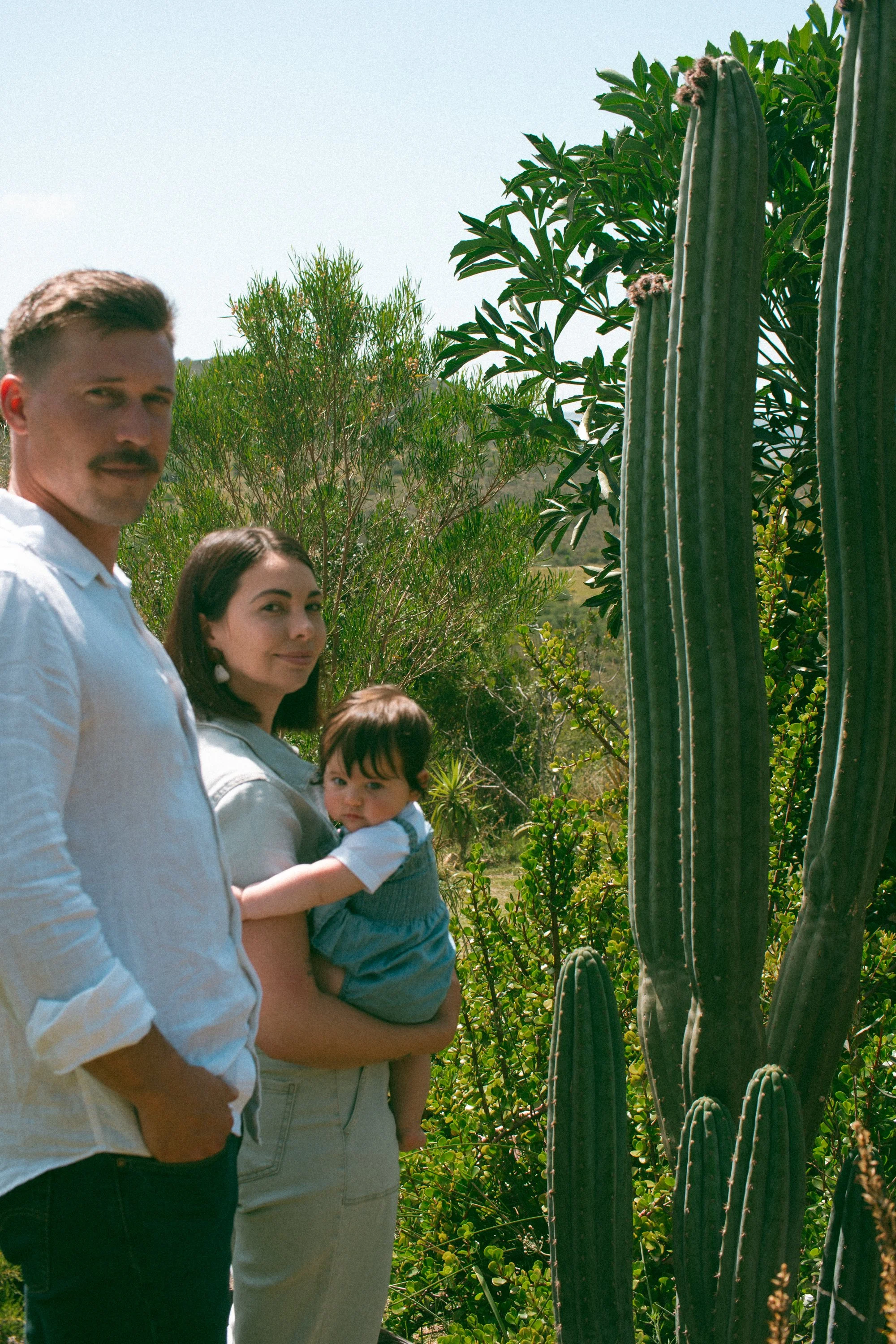 A family of three standing outdoors near large cactus plants with green leafy trees, under a clear sky on a sunny day.