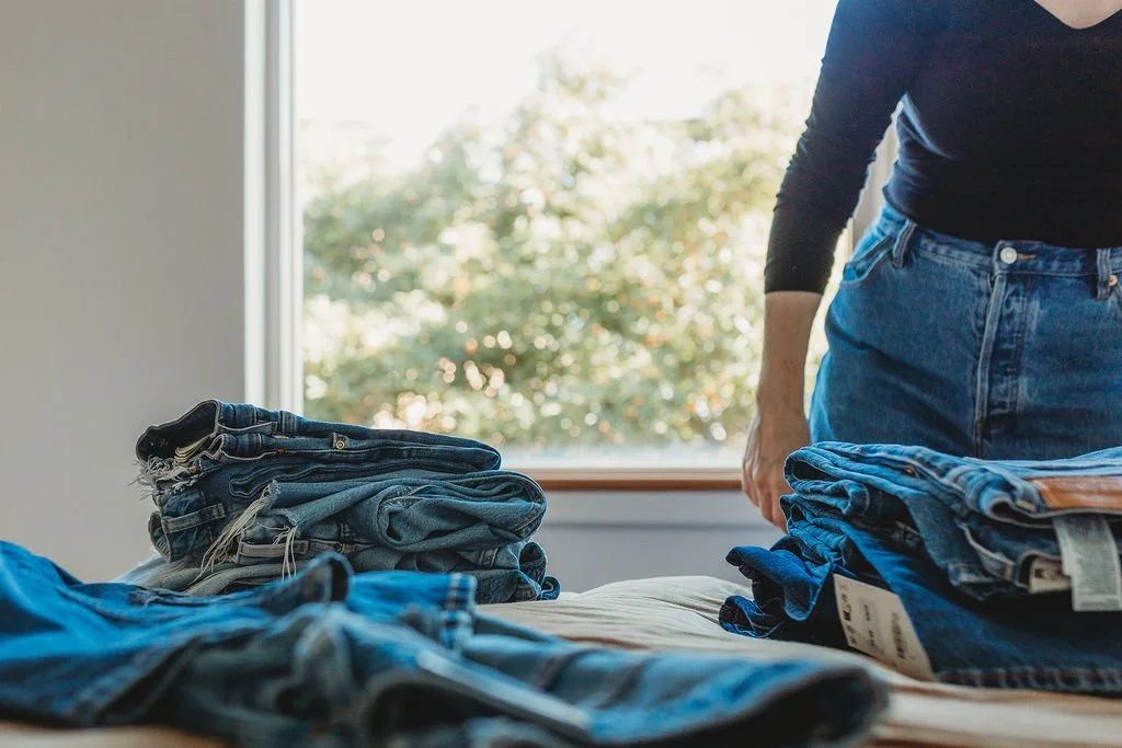 Person folding or sorting blue jeans on a bed near a window with tree outside.