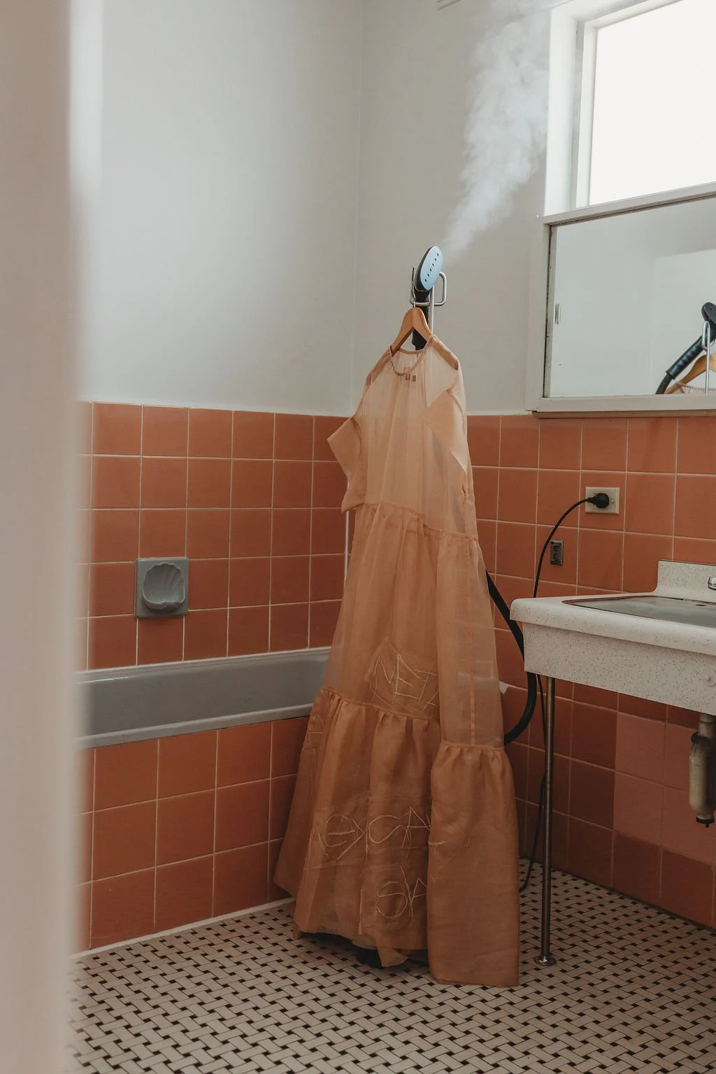 A vintage peach-colored dress hangs on a hanger over an ironing board in a laundry room, with steam rising from the iron.