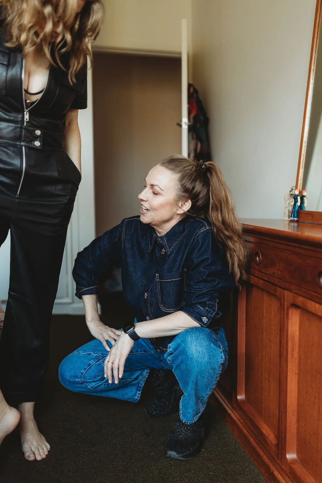 A woman with long brown hair in a ponytail wearing a dark denim shirt and jeans, squatting on the floor, talking to another woman wearing a black leather top and pants, who is standing barefoot.