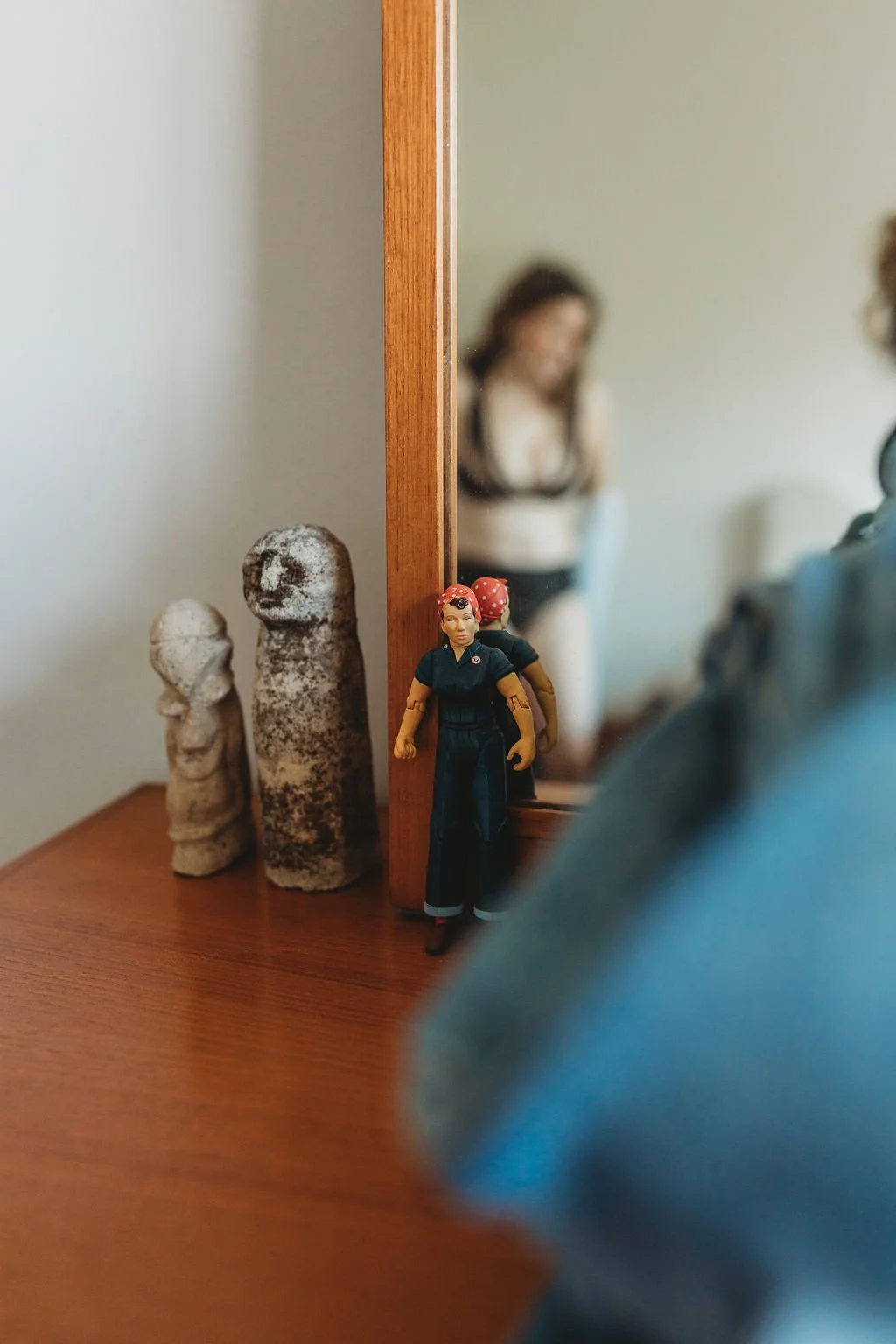 A wooden dresser with three stone carvings resembling heads and a toy firefighter figure standing in front of a mirror that reflects a woman.
