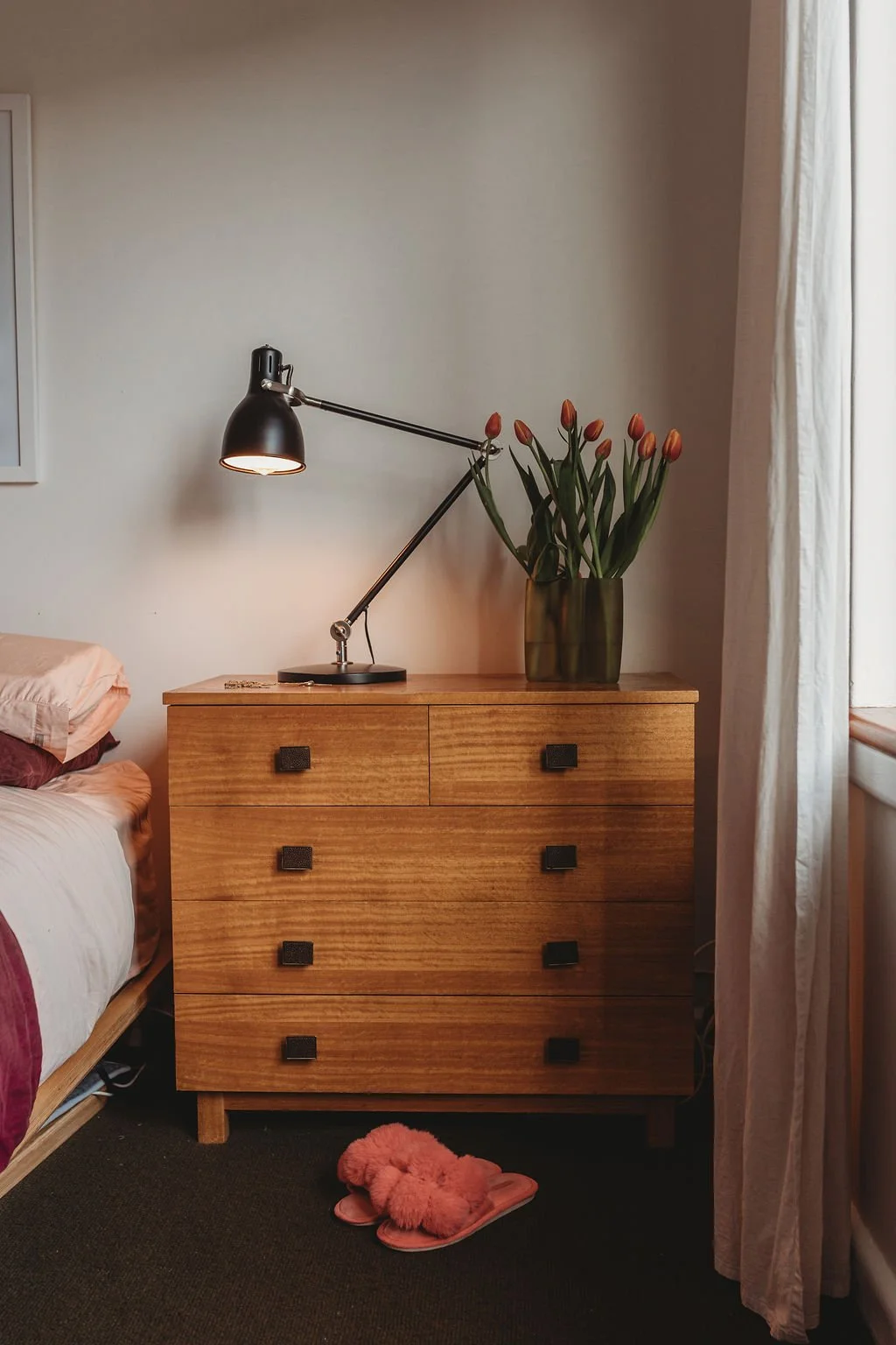 Wooden dresser with black square handles, topped with a green vase of pink tulips and a black adjustable desk lamp, in a room with a bed and beige curtains.