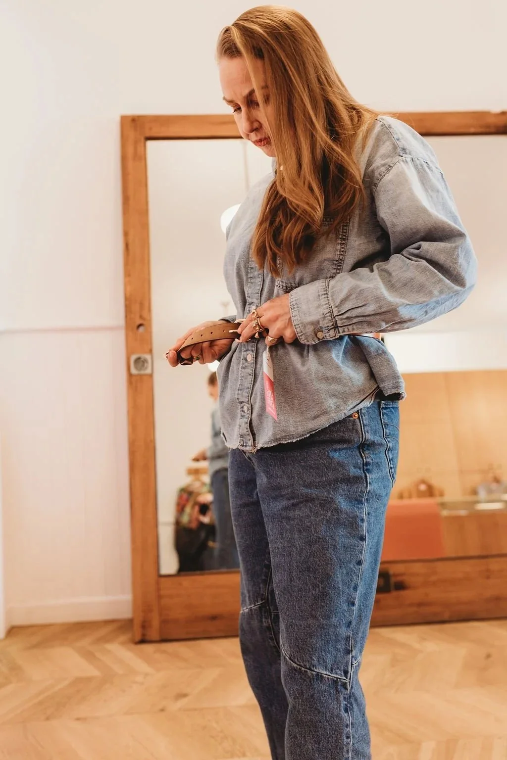 A woman with long red hair wearing a denim jacket and jeans looking at a brown belt in her hands, standing in front of a wooden framed mirror in a room with light-colored walls and wooden flooring.