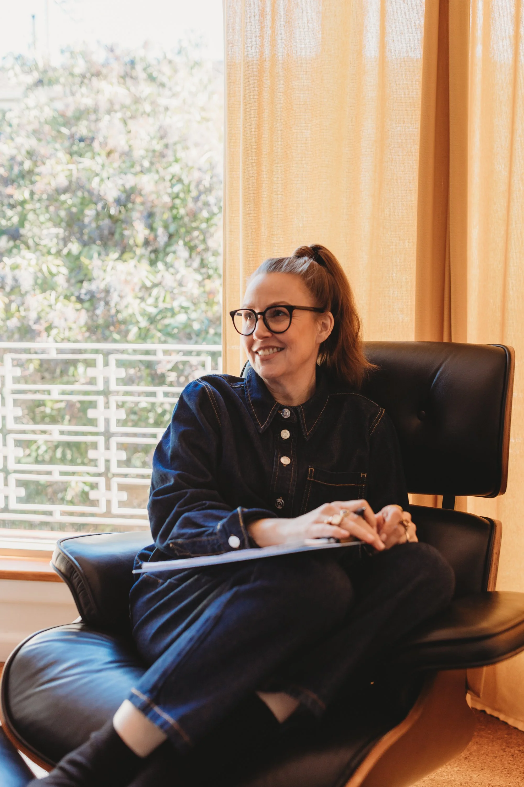A woman with glasses and red hair tied back, smiling while sitting on a black leather lounge chair near a window with yellow curtains.
