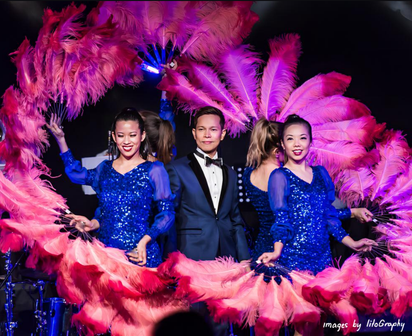 Group of performers, some women in glittery blue costumes with feathers, and a man in a tuxedo, on stage with a large pink feather fan backdrop.
